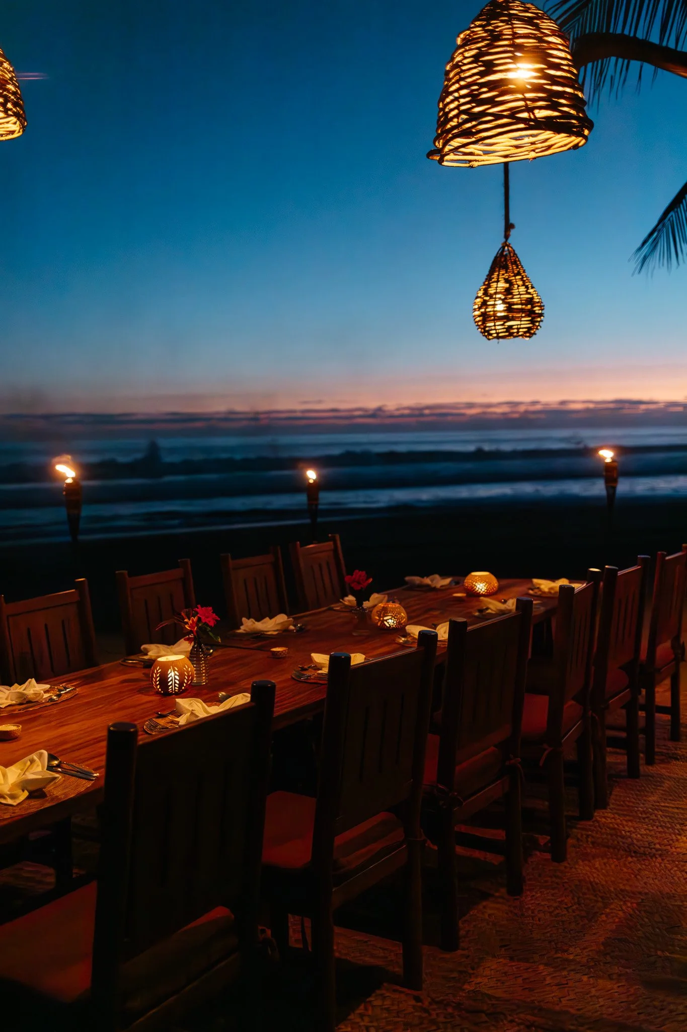 Outdoor dining area at sunset with a long wooden table, chairs, lit candles, and woven lantern pendant lights, overlooking the ocean.