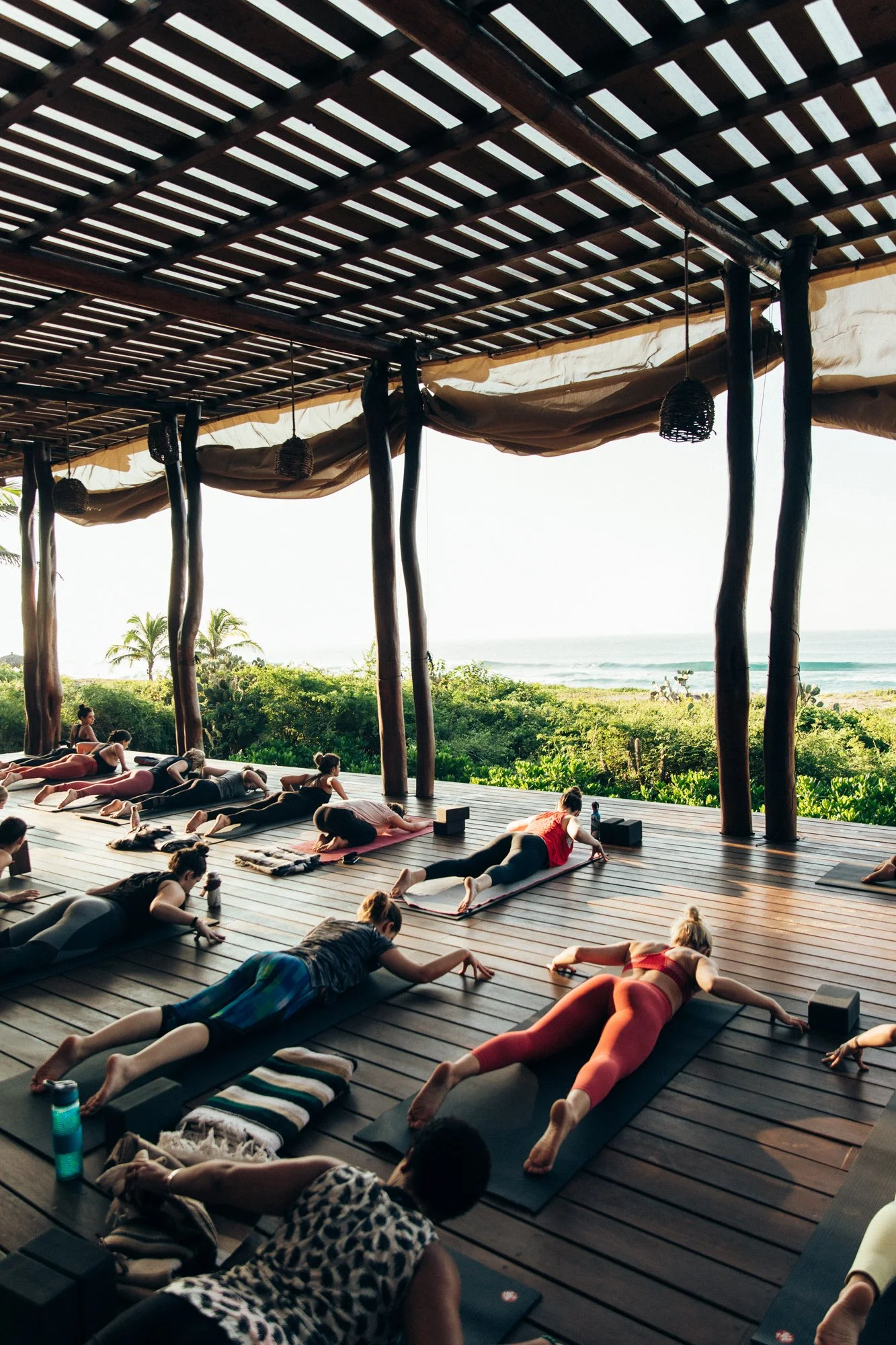Group of people practicing yoga outdoors on yoga mats on wooden platform overlooking greenery and ocean during daylight.