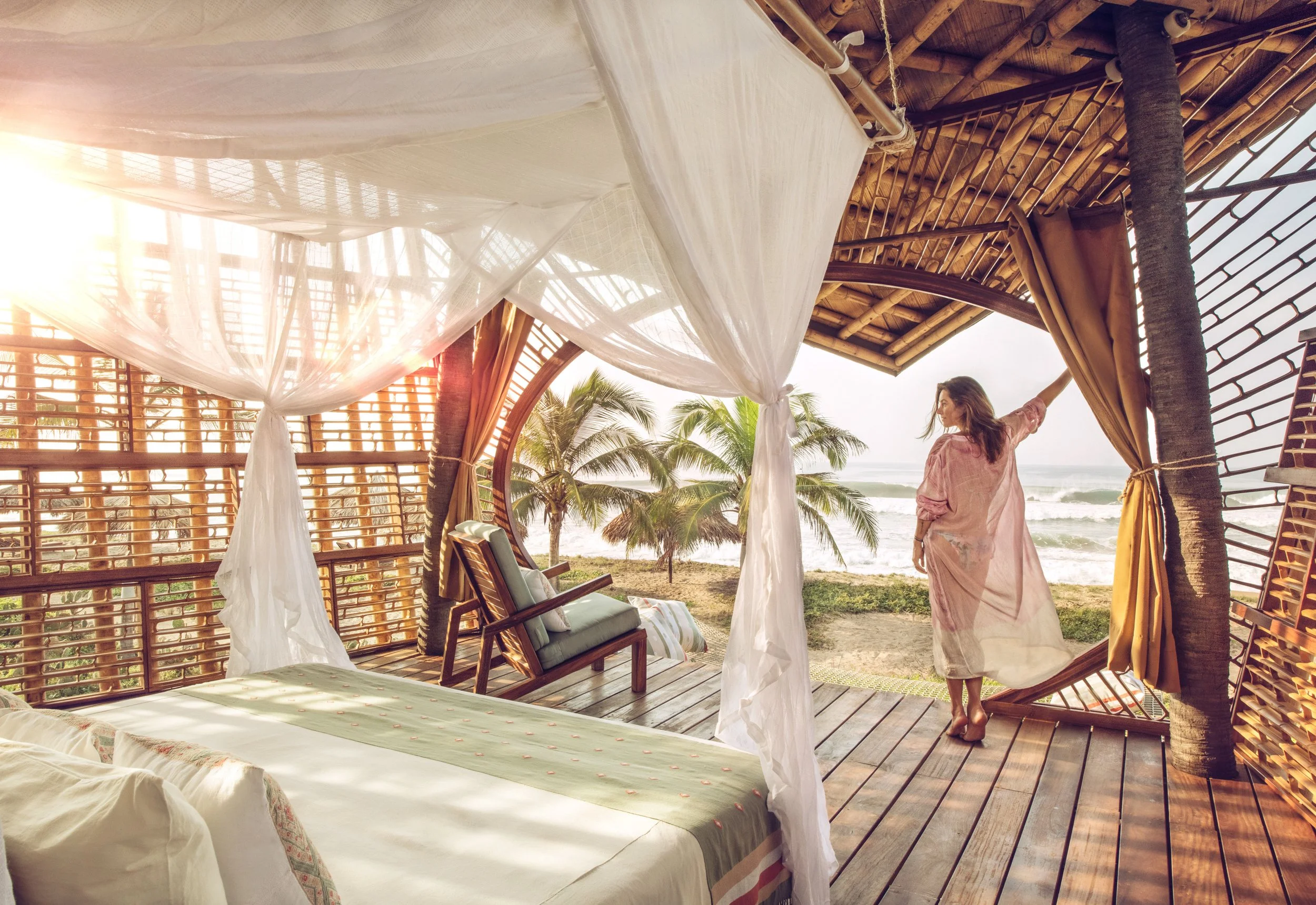 A woman in a pink dress standing on a wooden deck of a beachside hut, looking out at palm trees and the ocean during sunset.