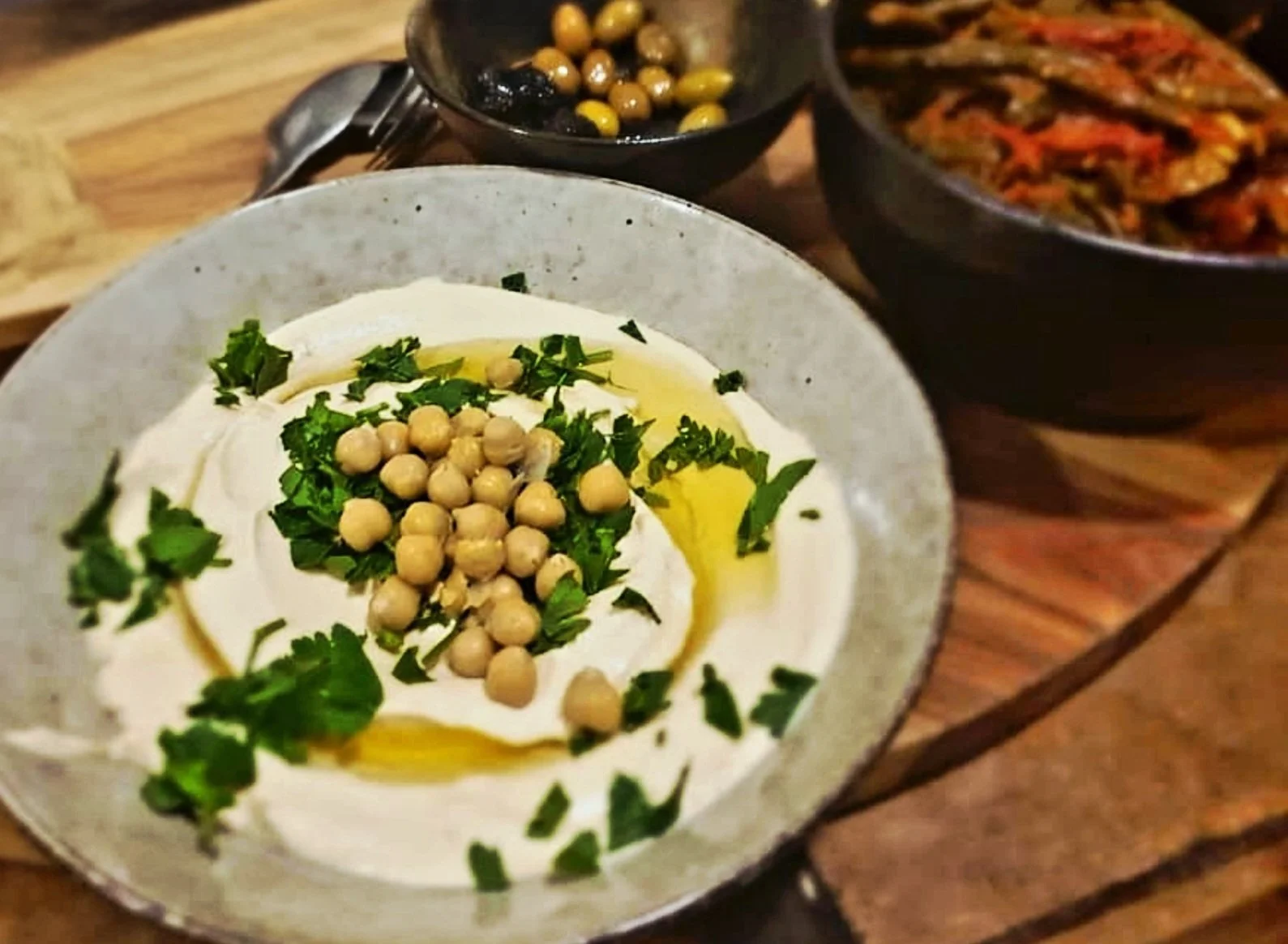 A bowl of creamy hummus topped with chickpeas, chopped herbs, and olive oil. In the background, two bowls contain cooked chickpeas and roasted vegetables.