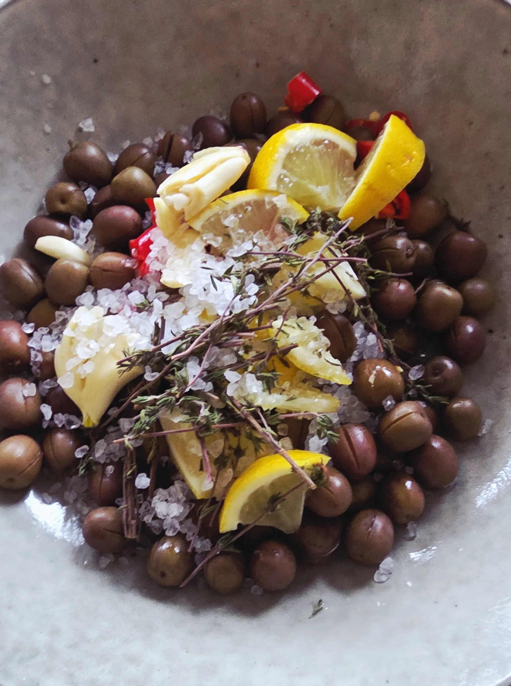 A mixture of sliced lemons, red chili peppers, fresh thyme sprigs, and brined olives on crushed ice in a metallic container.