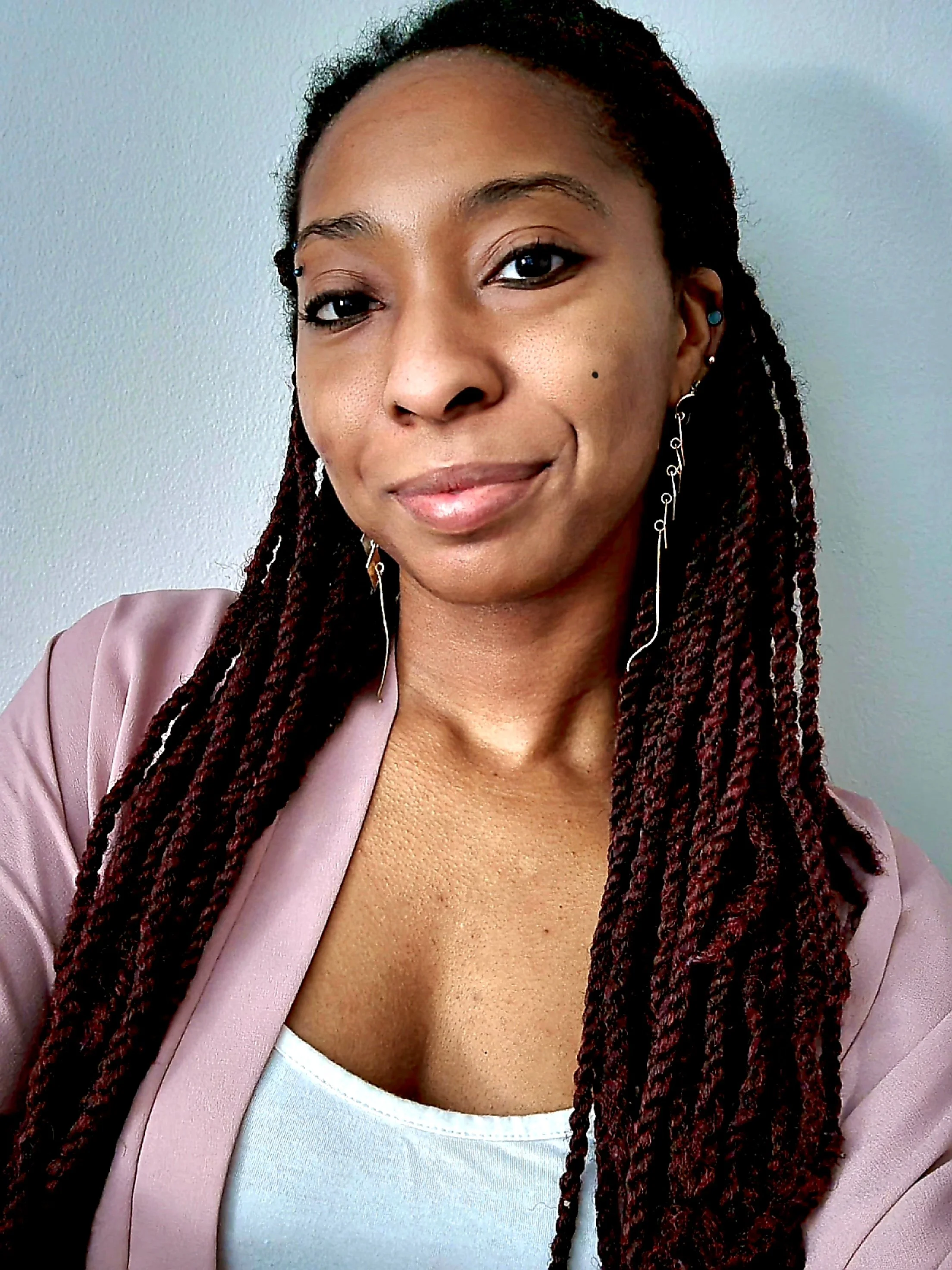 A close-up of a woman with long braided hair, wearing earrings and a light pink blazer, standing against a plain background.