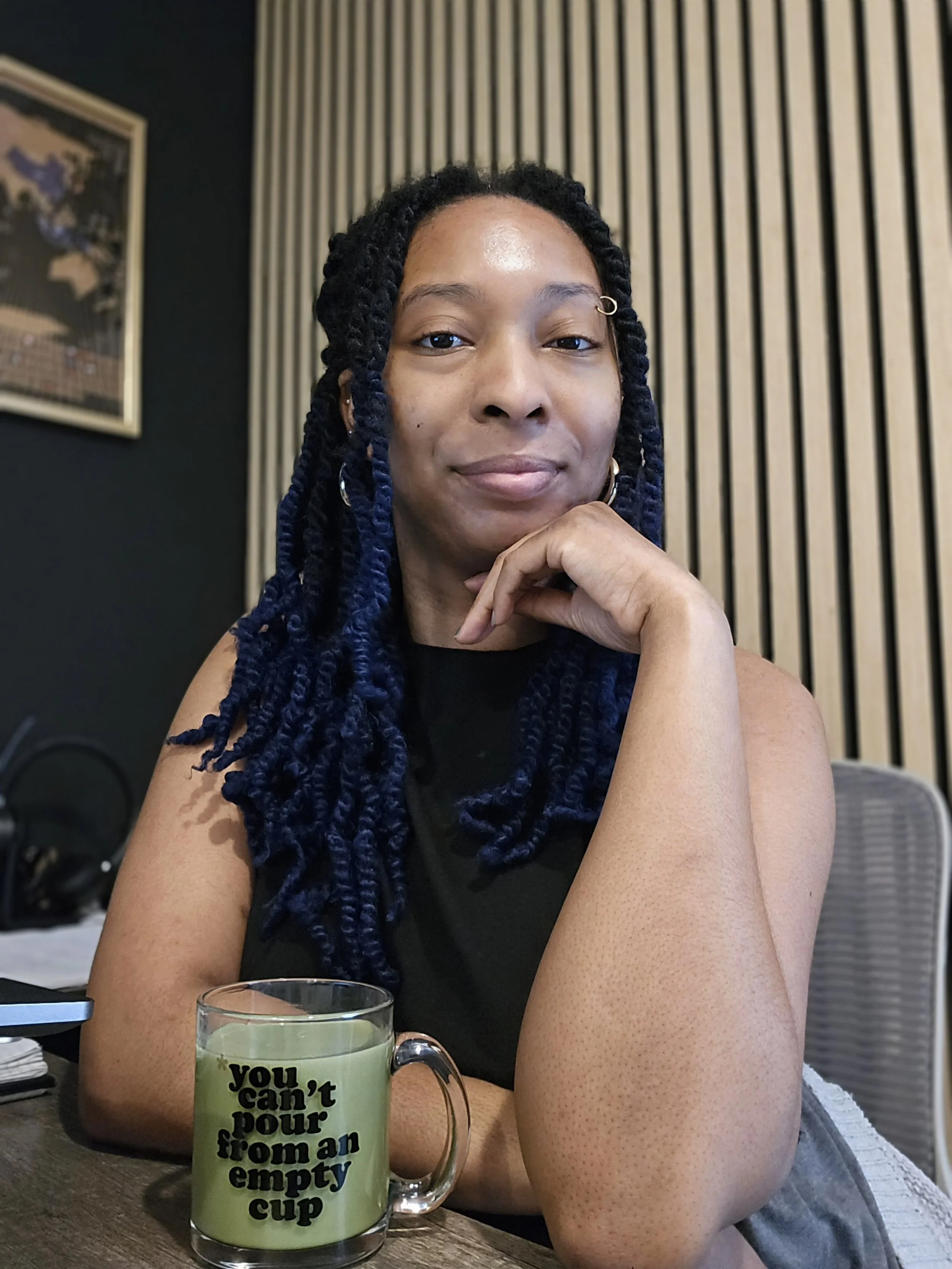 picture of a Black woman resting arm on table and one hand under chin, sitting at a desk, with a mug that says "you can't pour from an empty cup"