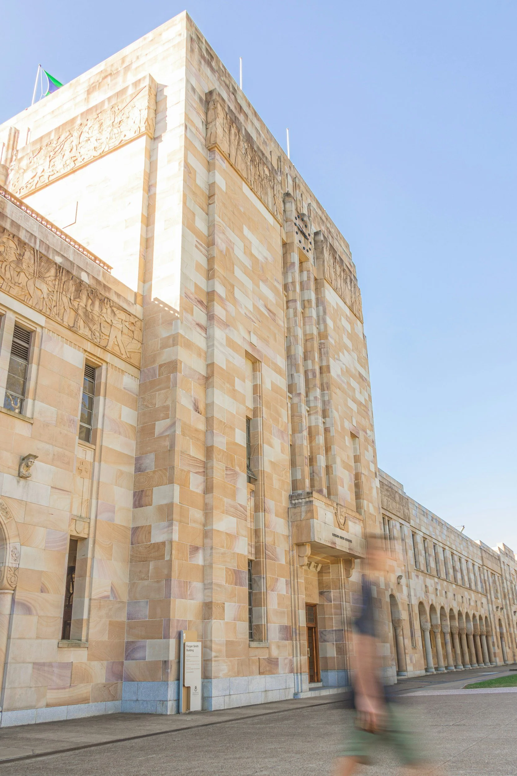 A tall, tan brick building with intricate carvings and sculptures, featuring multiple windows and a flag on top, against a clear blue sky. There is a person walking past in front of the building.