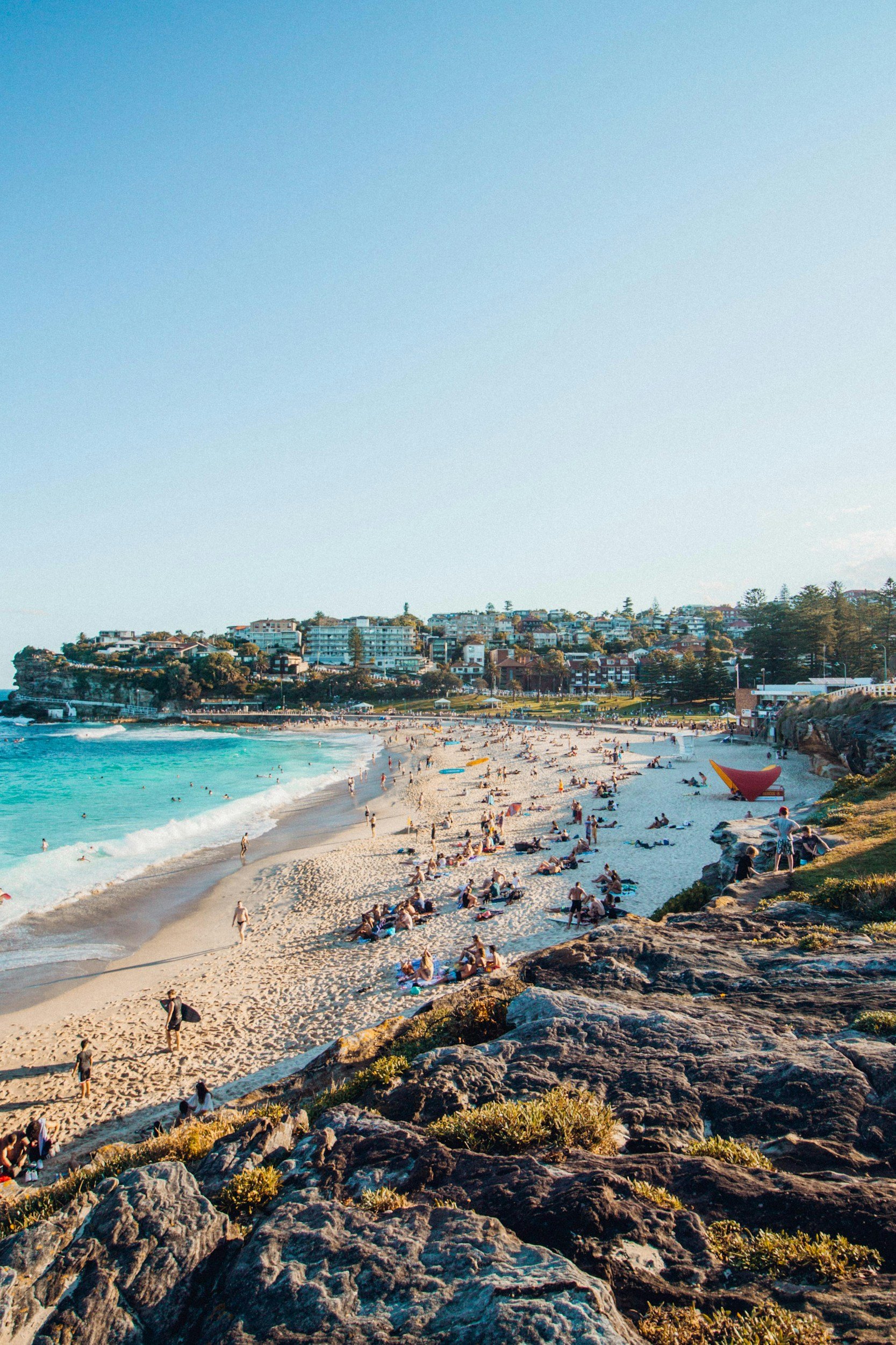 Beach with many people relaxing and swimming, ocean waves, rocky foreground, buildings on a hill in the background, clear blue sky.
