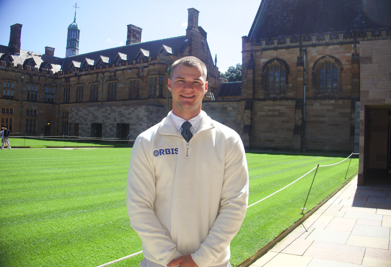 Young man in an off-white jacket standing outdoors on a sunny day in front of a historic stone building with large windows and a well-maintained lawn.