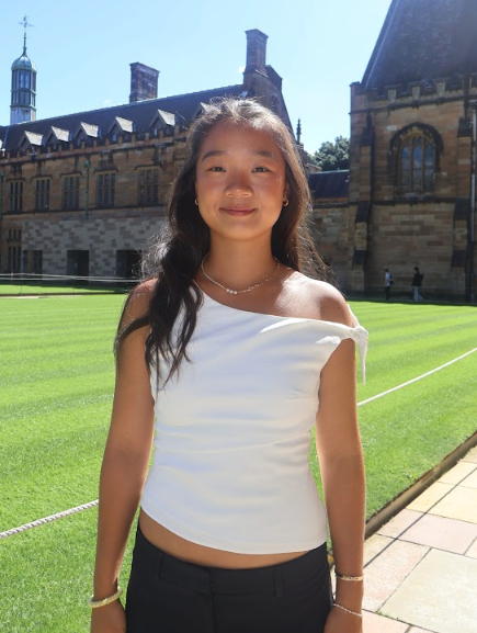 Young girl standing outdoors in front of a historic stone building with a manicured lawn and a pathway, smiling at the camera.