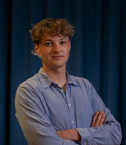 Young man with curly hair wearing a striped shirt, standing with arms crossed in front of a blue curtain.