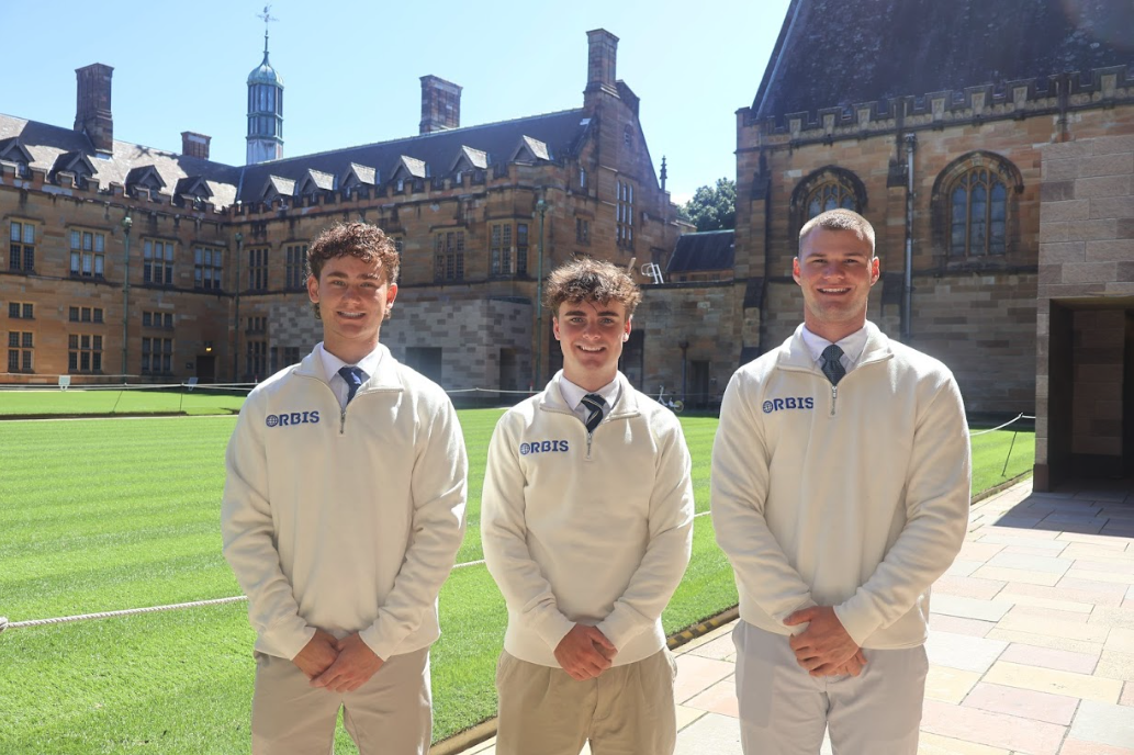 Three young men in white sweaters with 'ORBIS' logo standing outdoors in front of a historic stone building with a well-manicured grass lawn, on a sunny day.