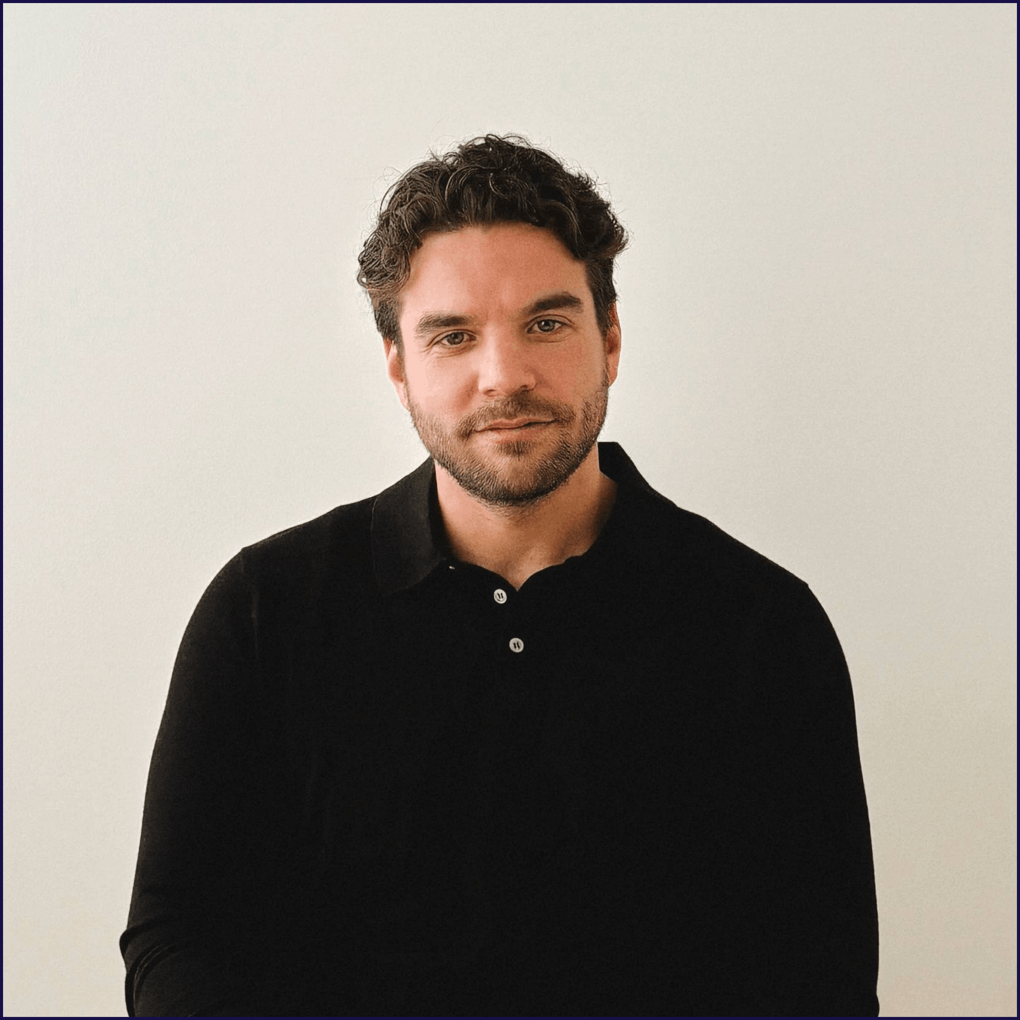 Headshot of a young man with dark wavy hair, a beard, wearing a black polo shirt, standing against a plain white background.