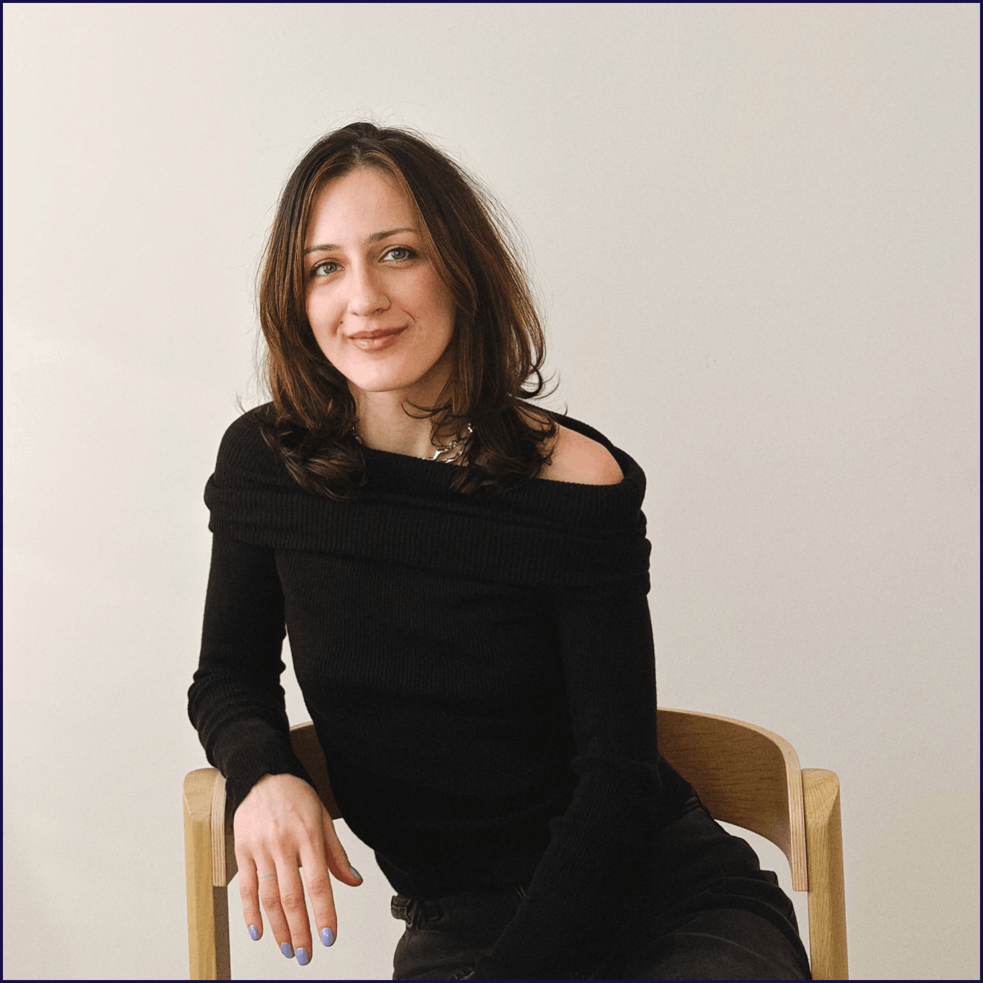 A woman sitting on a wooden chair with her left hand resting on the chair's armrest. She has shoulder-length brown hair, blue eyes, and is smiling slightly. She is wearing a black off-shoulder top and has painted nails. The background is plain and light-colored.