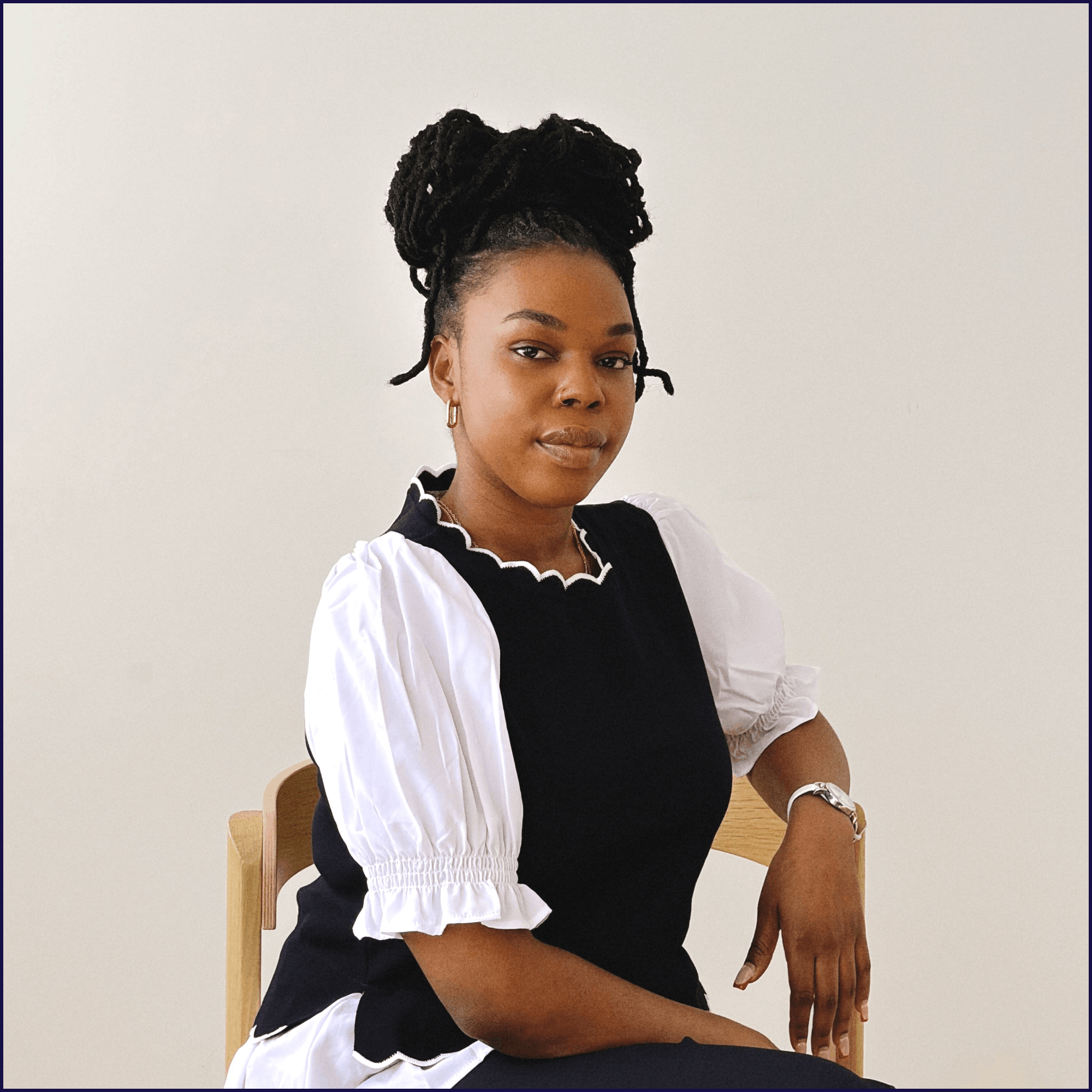 A woman with dark skin and styled dreadlocks sitting on a wooden chair against a plain light-colored background. She is wearing a black and white top with puffed sleeves and has a confident expression.