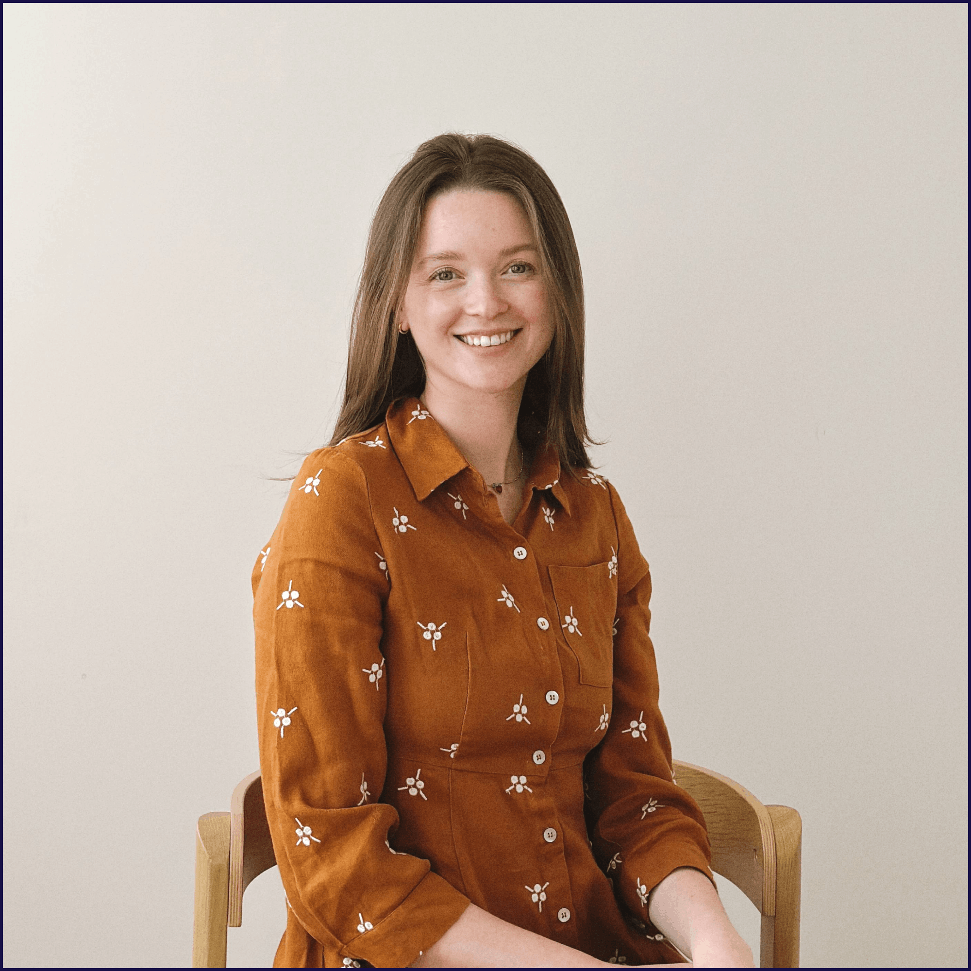 A woman with long brown hair smiling while sitting on a wooden chair against a plain white wall, wearing a brown shirt with white flower embroidery.