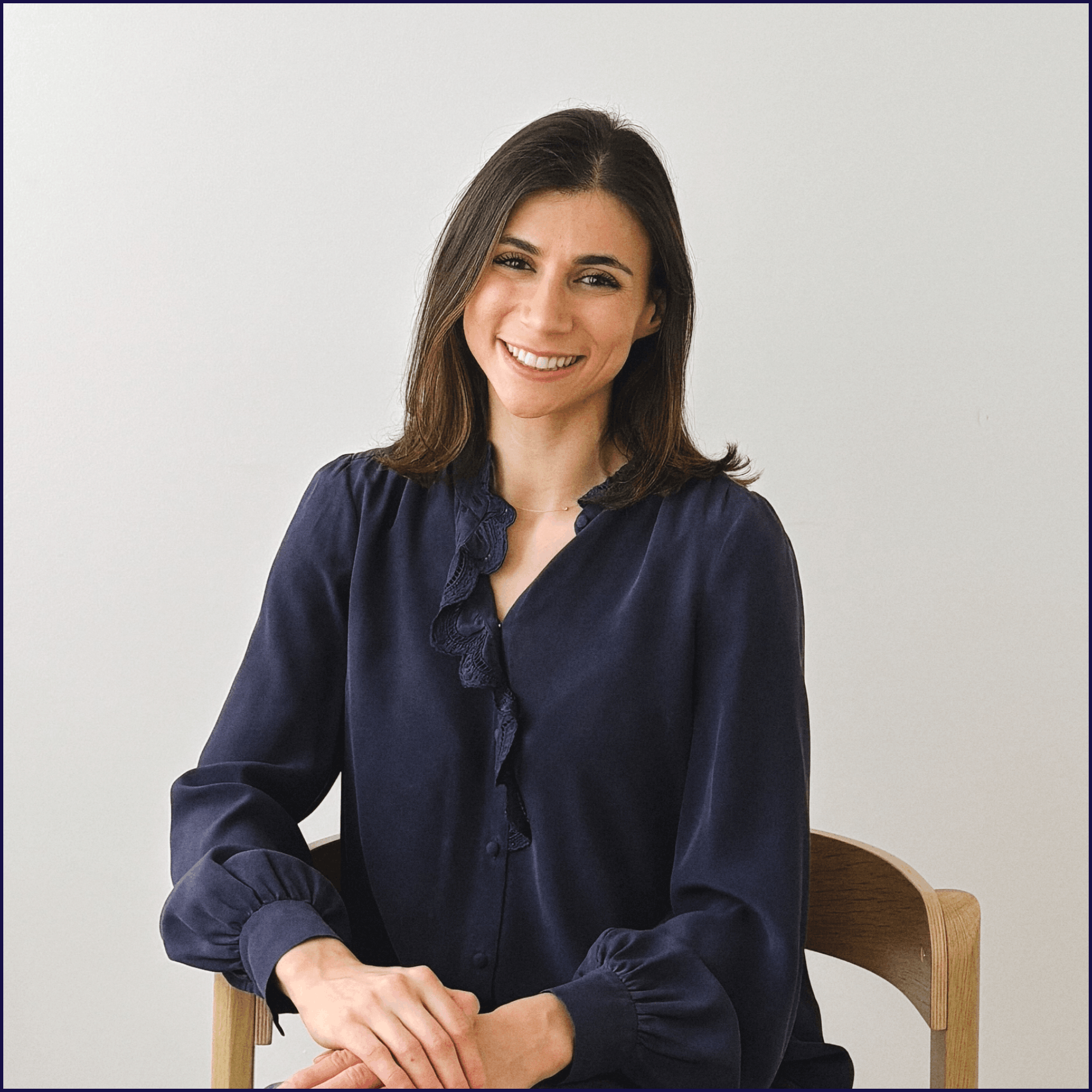 A woman with shoulder-length brown hair smiling while sitting at a wooden table against a plain white wall, wearing a dark blue blouse with ruffled details.