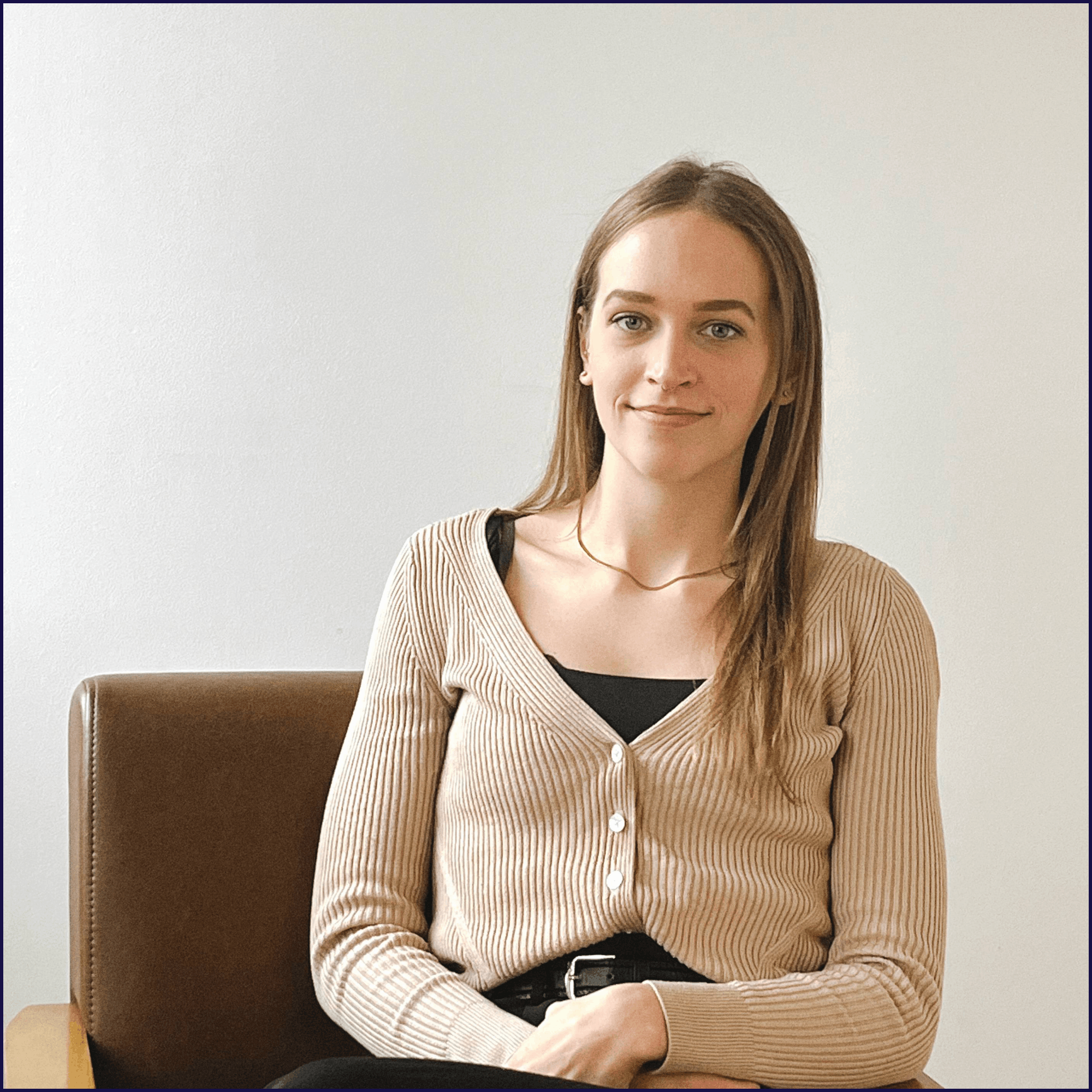 A young woman with long, light brown hair, blue eyes, and fair skin sitting on a brown leather chair against a plain white wall. She is wearing a beige ribbed cardigan over a black top and has a subtle smile.