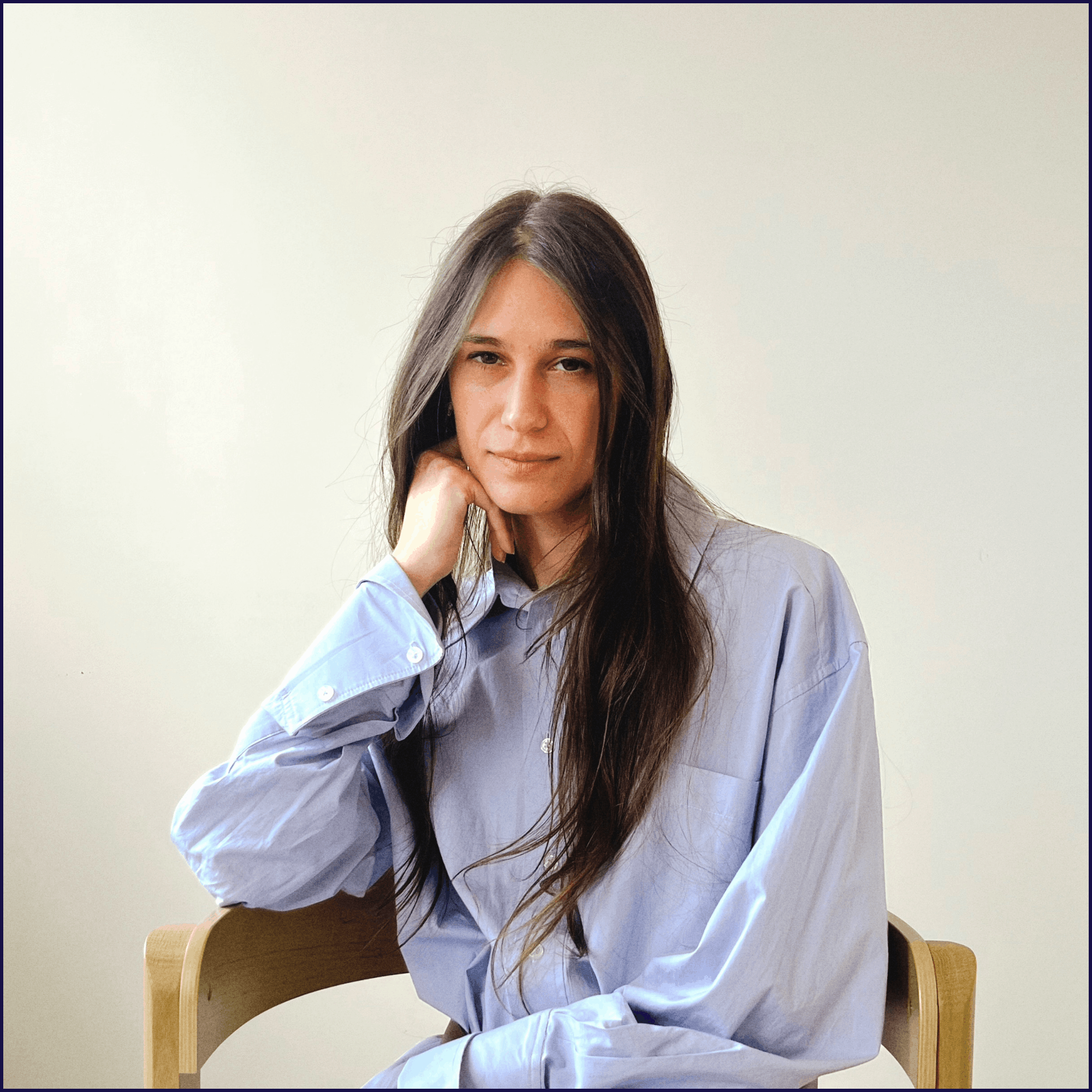 A woman with long dark hair and a calm, neutral expression, seated on a wooden chair against a plain cream-coloured background.