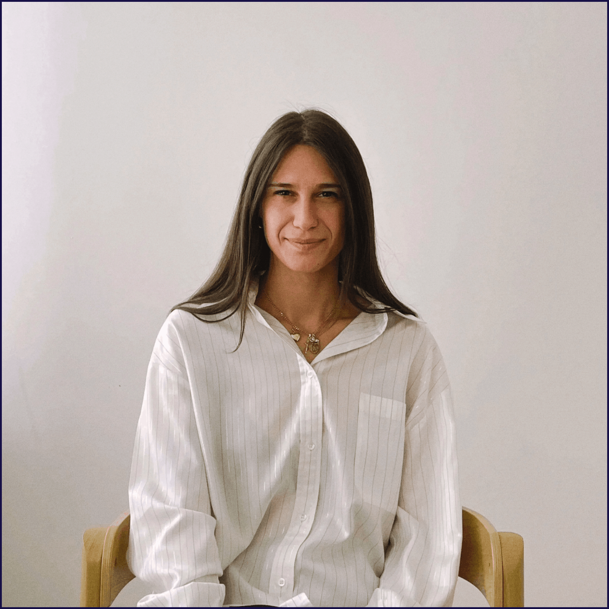 A woman with long dark hair and a calm, neutral expression, seated on a wooden chair against a plain cream-coloured background.