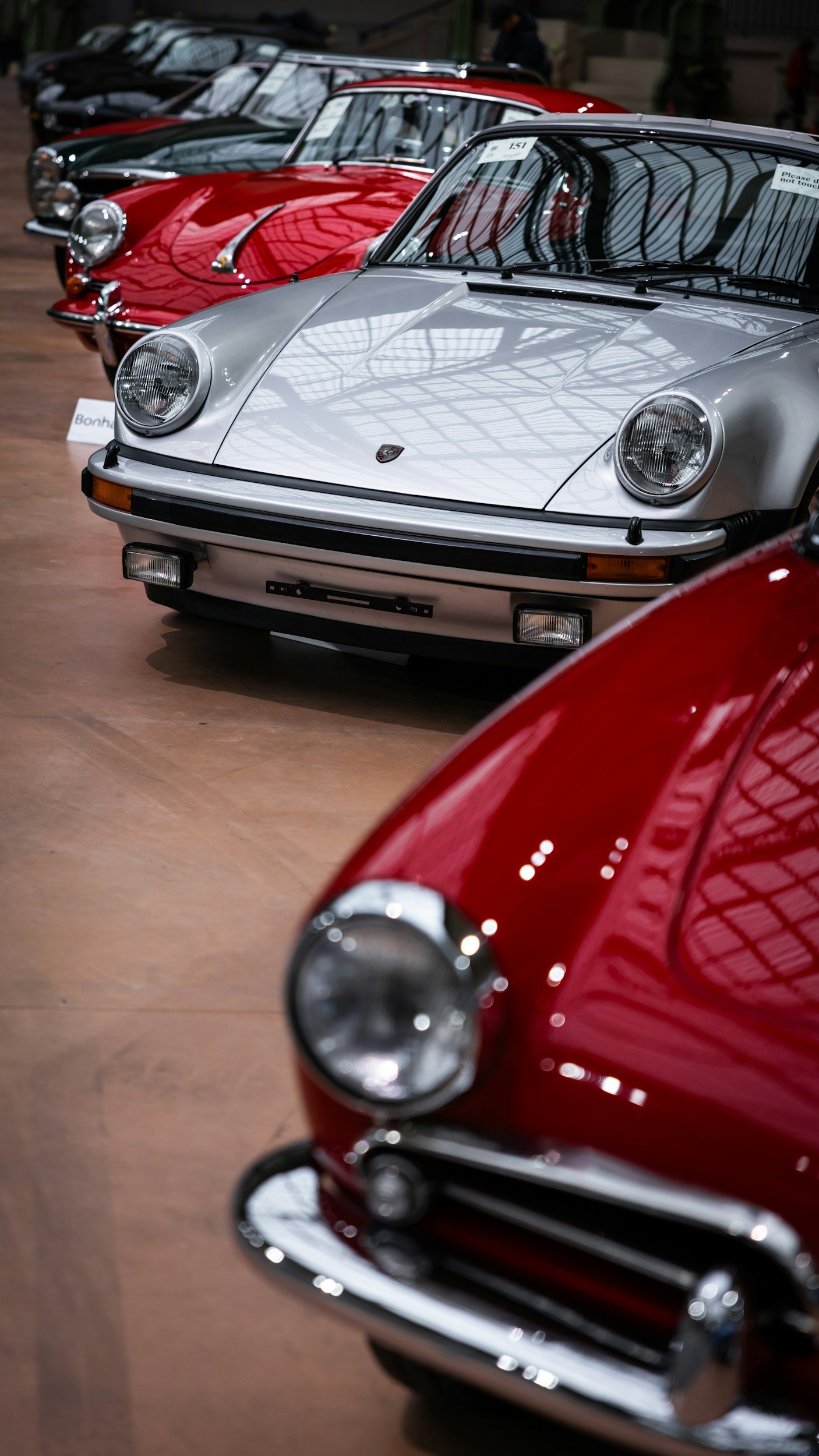 Lineup of vintage sports cars in an exhibition hall, including a silver Porsche 911 in the foreground, with red and black cars visible behind it.