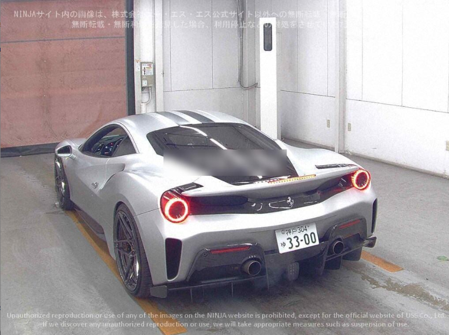 A silver sports car parked indoors, facing away. It has circular brake lights and dual exhaust pipes, with a black rear wing and tinted windows.