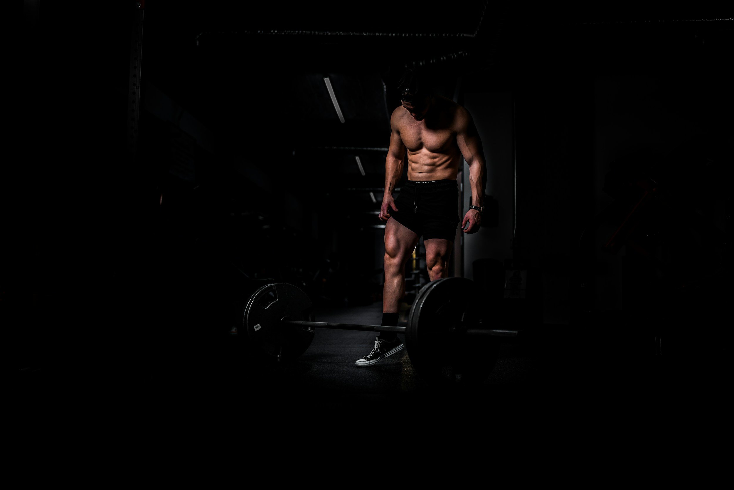 A shirtless man wearing black shorts and sneakers standing in a dark gym, looking down at a barbell on the floor.