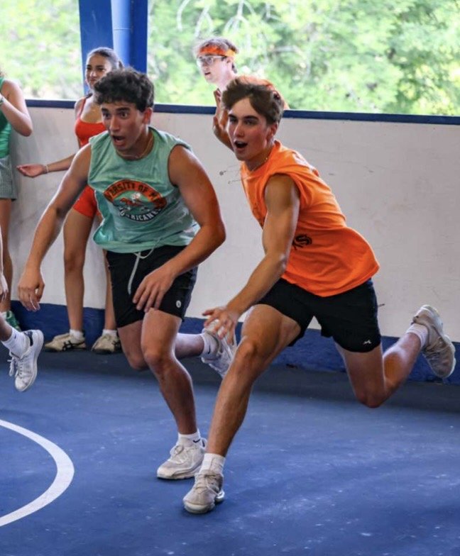Two boys in athletic clothing are sprinting on an indoor basketball court, with other children standing in the background by a wall and large window. One boy in a green tank top and black shorts looks surprised, while the boy in an orange shirt and black shorts is focused on running.