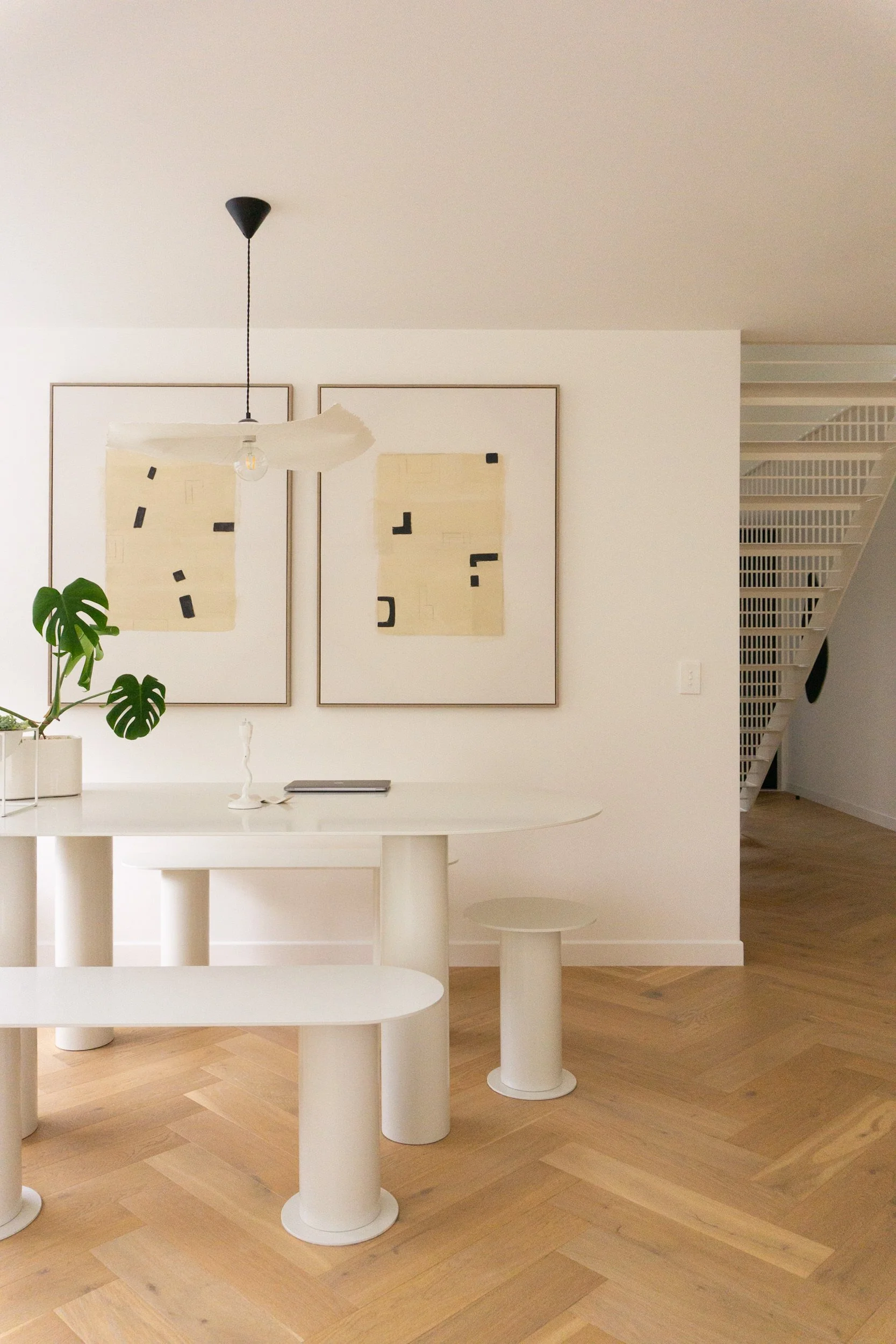 Minimalist white dining area with white table, stool, and potted Monstera plant, contemporary artwork on white walls, wooden floor, modern lighting fixture.
