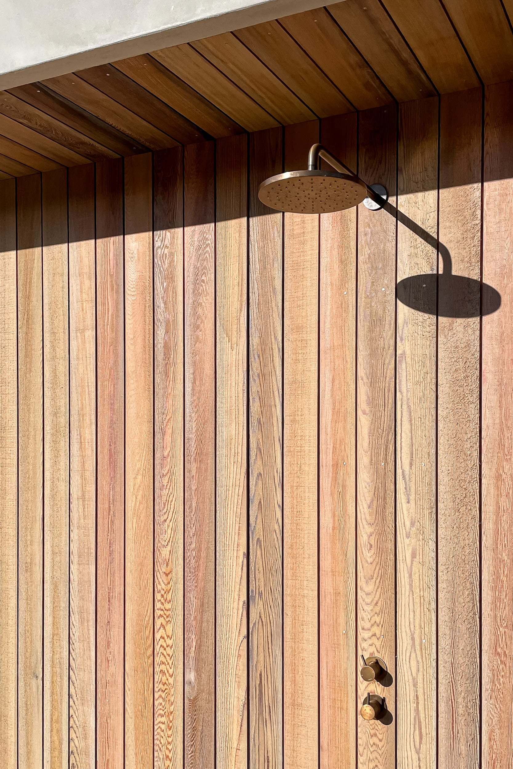 Outdoor shower with a showerhead mounted on a wooden wall, with a shadow cast by the showerhead, and a bronze-colored control knob below it.