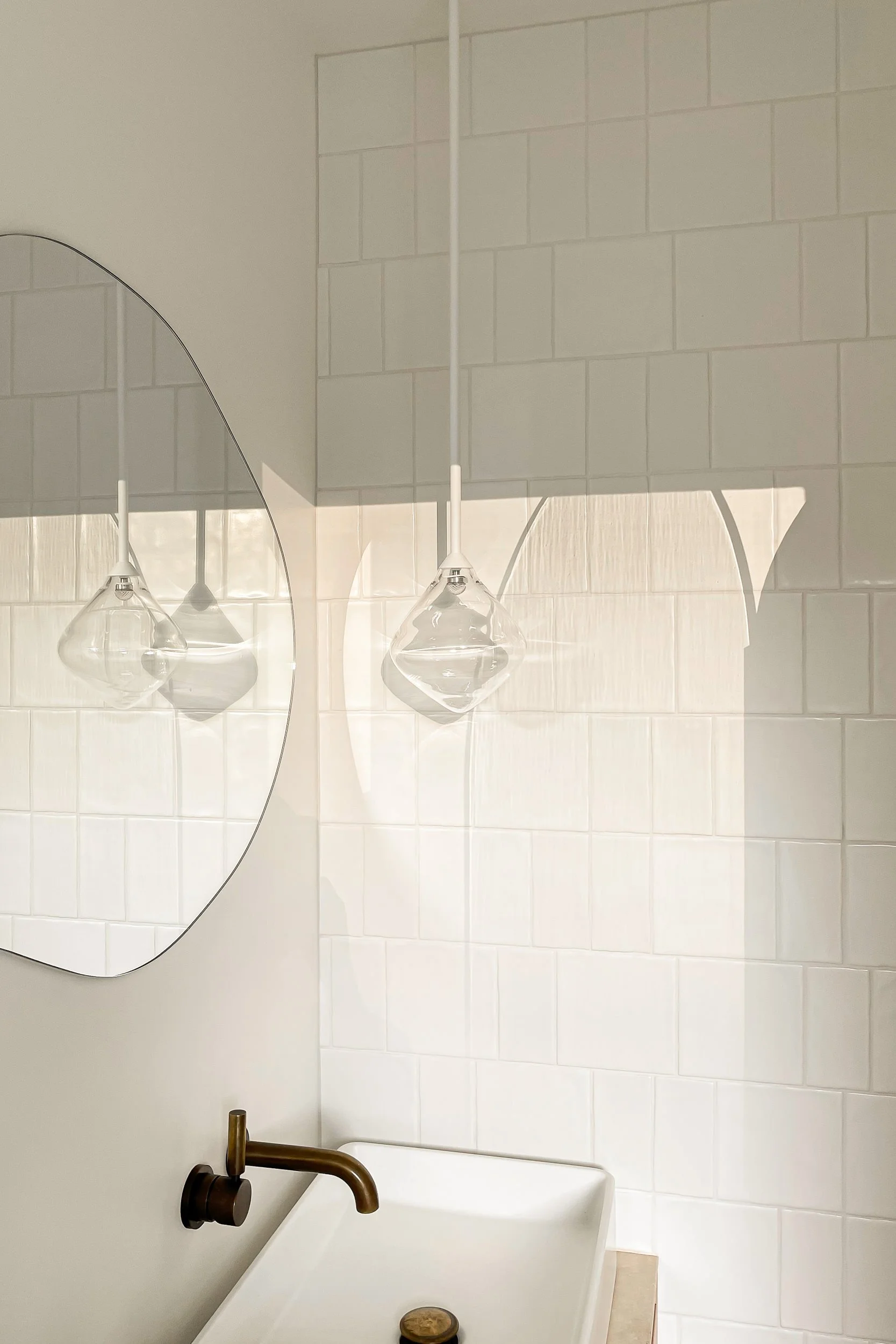 A bathroom sink with a bronze faucet and a round soap dish on the edge, with a large round mirror reflecting two hanging pendant lights with glass shades and a white tiled wall in the background.