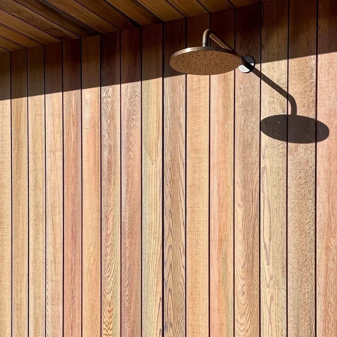 An outdoor shower with a rain-style showerhead mounted on a wooden wall, casting a shadow in bright sunlight.