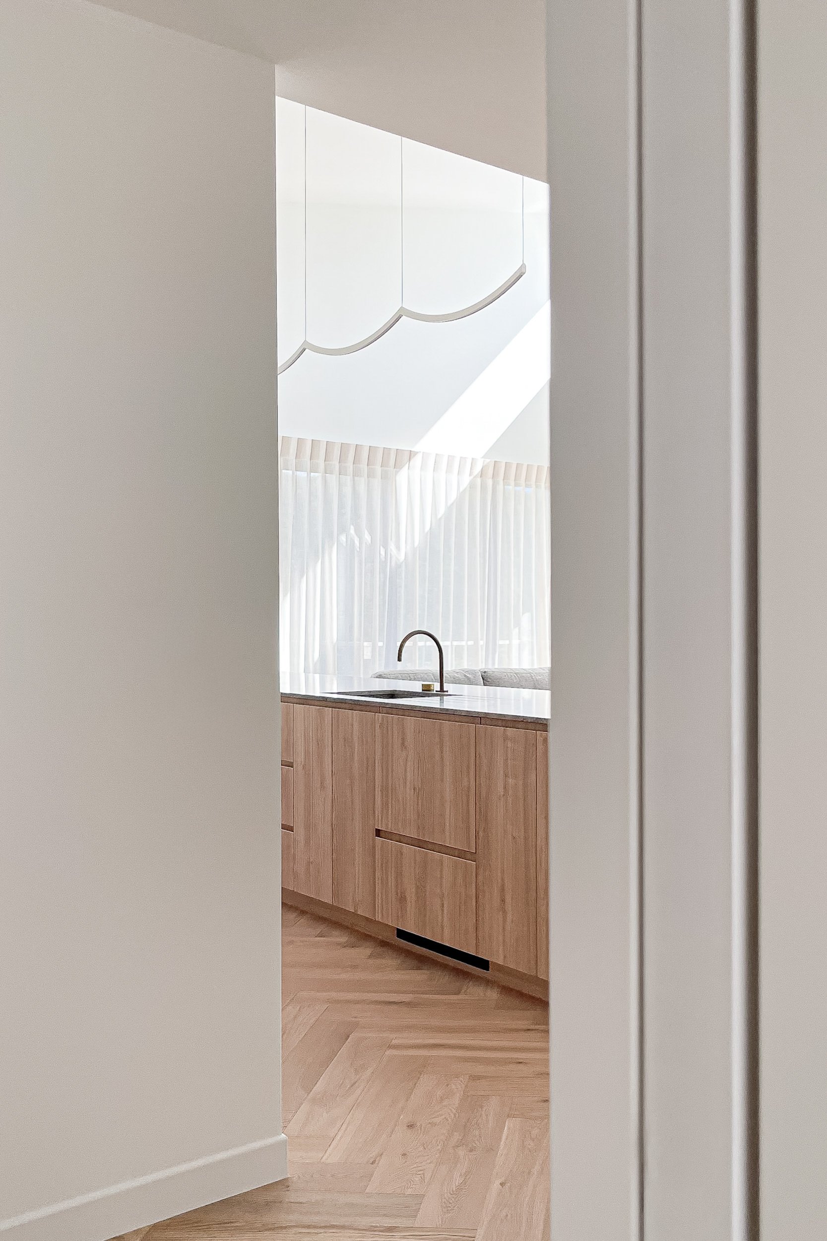 View of a modern kitchen with wooden cabinetry, a black faucet, and a bright window with sheer curtains, seen through a partially open white door.