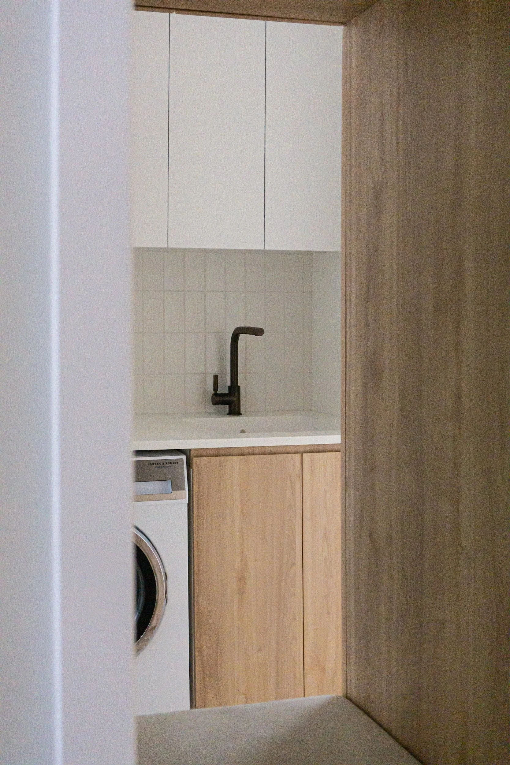 A kitchen with white upper cabinets, a black faucet, a beige tiled backsplash, lower beige cabinets, and a white washing machine.