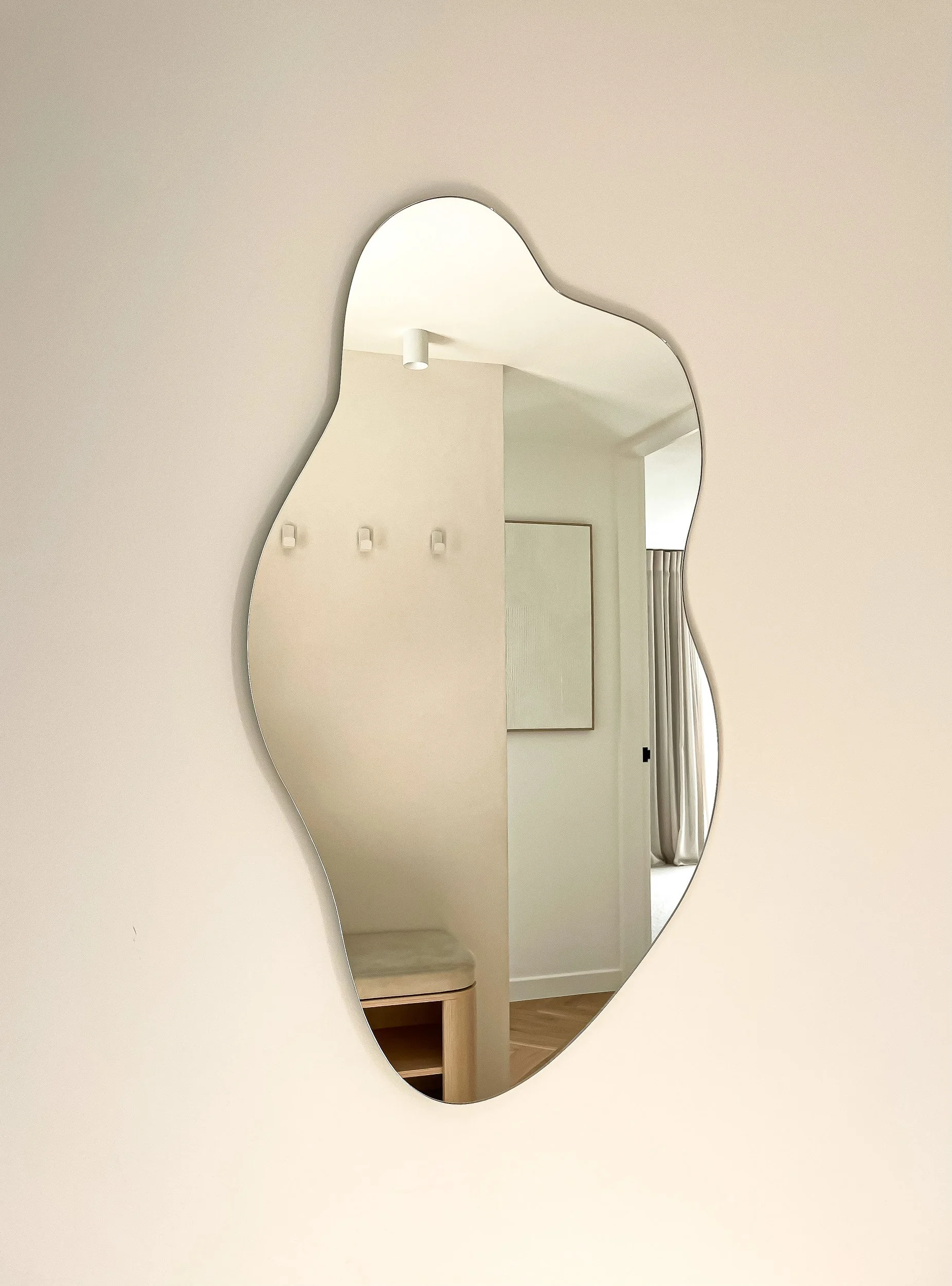 A reflection in a uniquely shaped mirror shows part of a beige room with wall hooks, a door frame, a curtain, and a glimpse of a wooden floor.