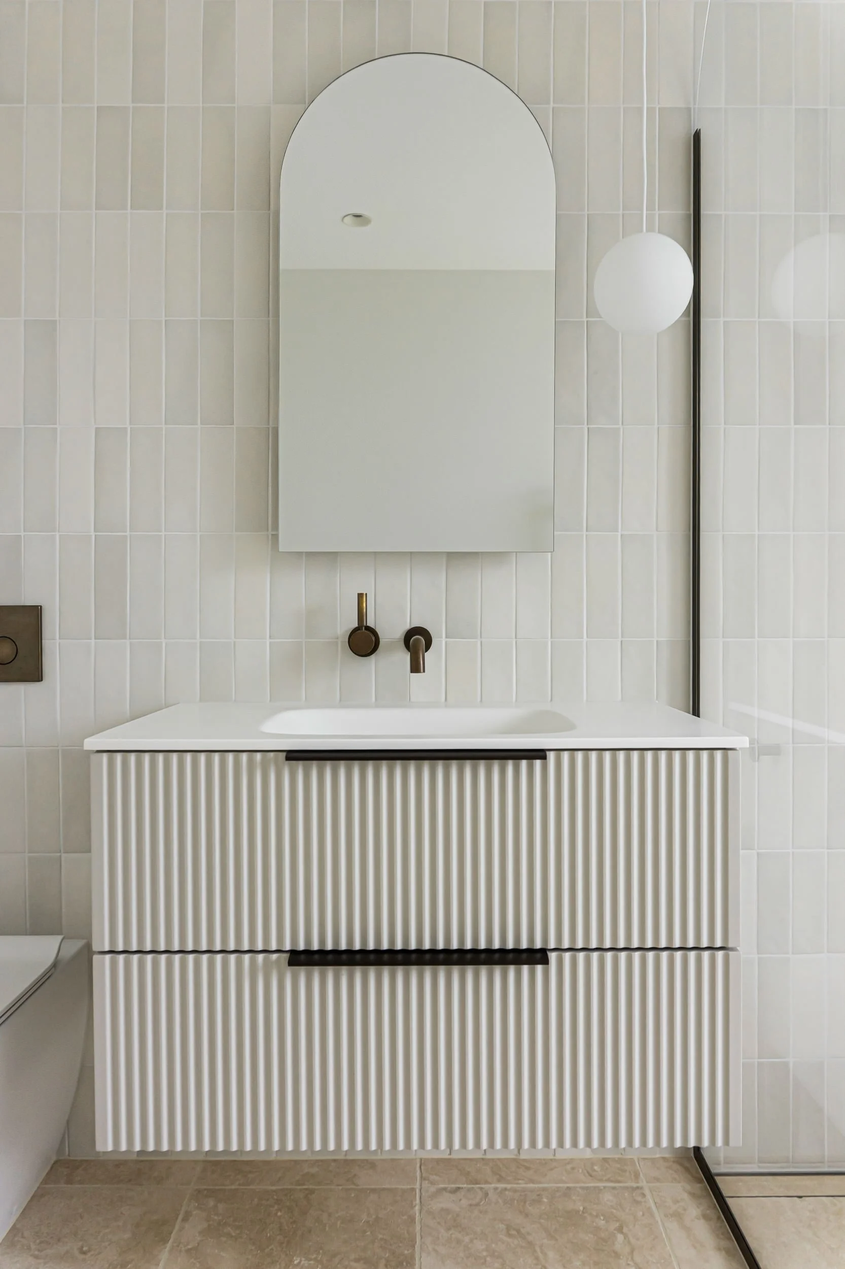 Modern bathroom with walk-in shower, oval bathtub, beige tiled walls and floor, oval mirror, minimalist vanity, black fixtures, glass divider, and pendant lighting.