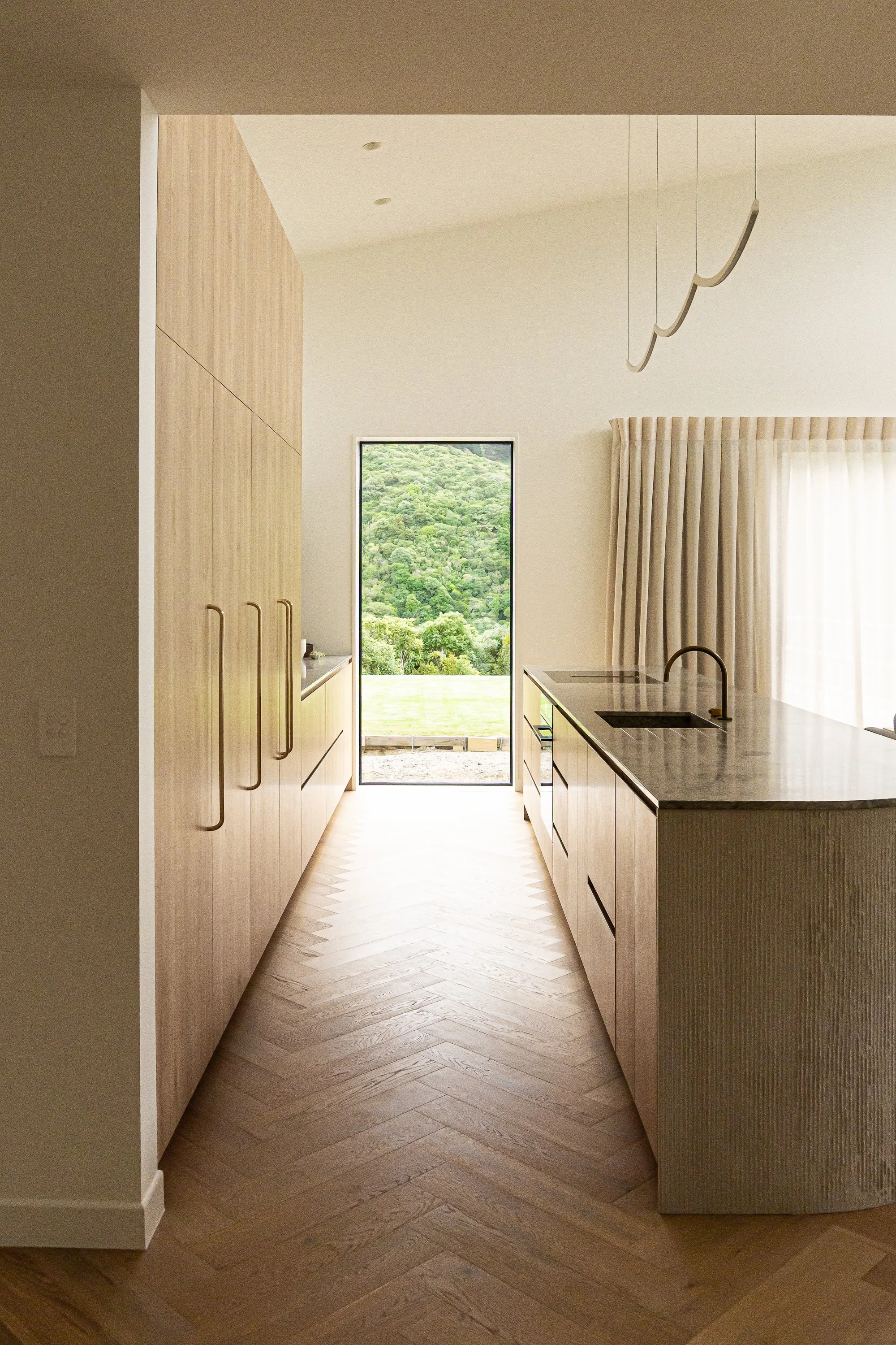 Bright, modern kitchen with a curved island, light wood cabinets, bar stools, and a window showing greenery outside.
