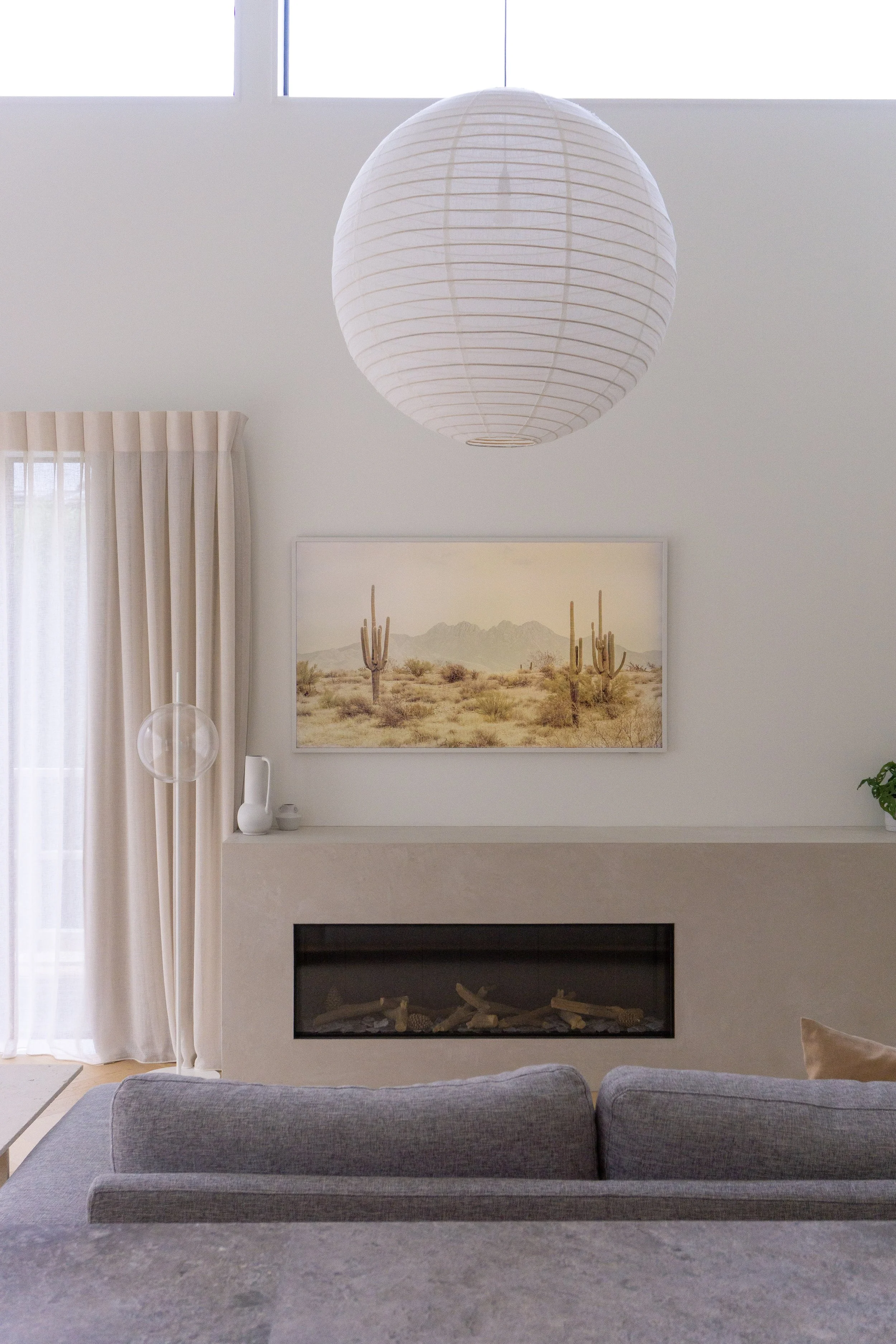 Living room with a large white paper lantern hanging from the ceiling, a landscape painting of desert cacti and mountains above a fireplace, gray sofa in foreground, and light-colored curtains next to a window.