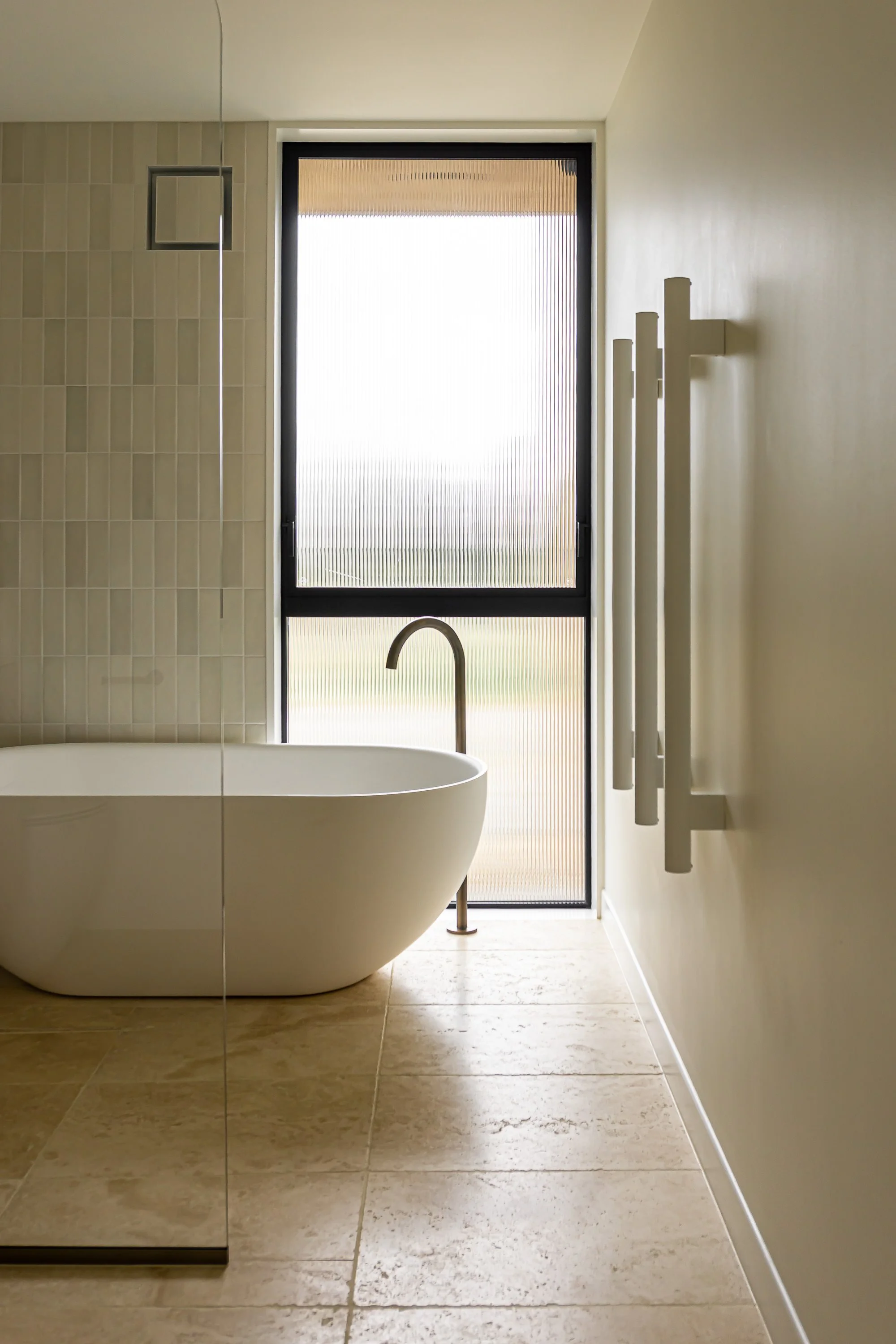 A modern bathroom with a floating white bathtub, beige tiled floor, and a large window with frosted glass, allowing natural light to enter. A set of vertical towel bars is mounted on the right wall.