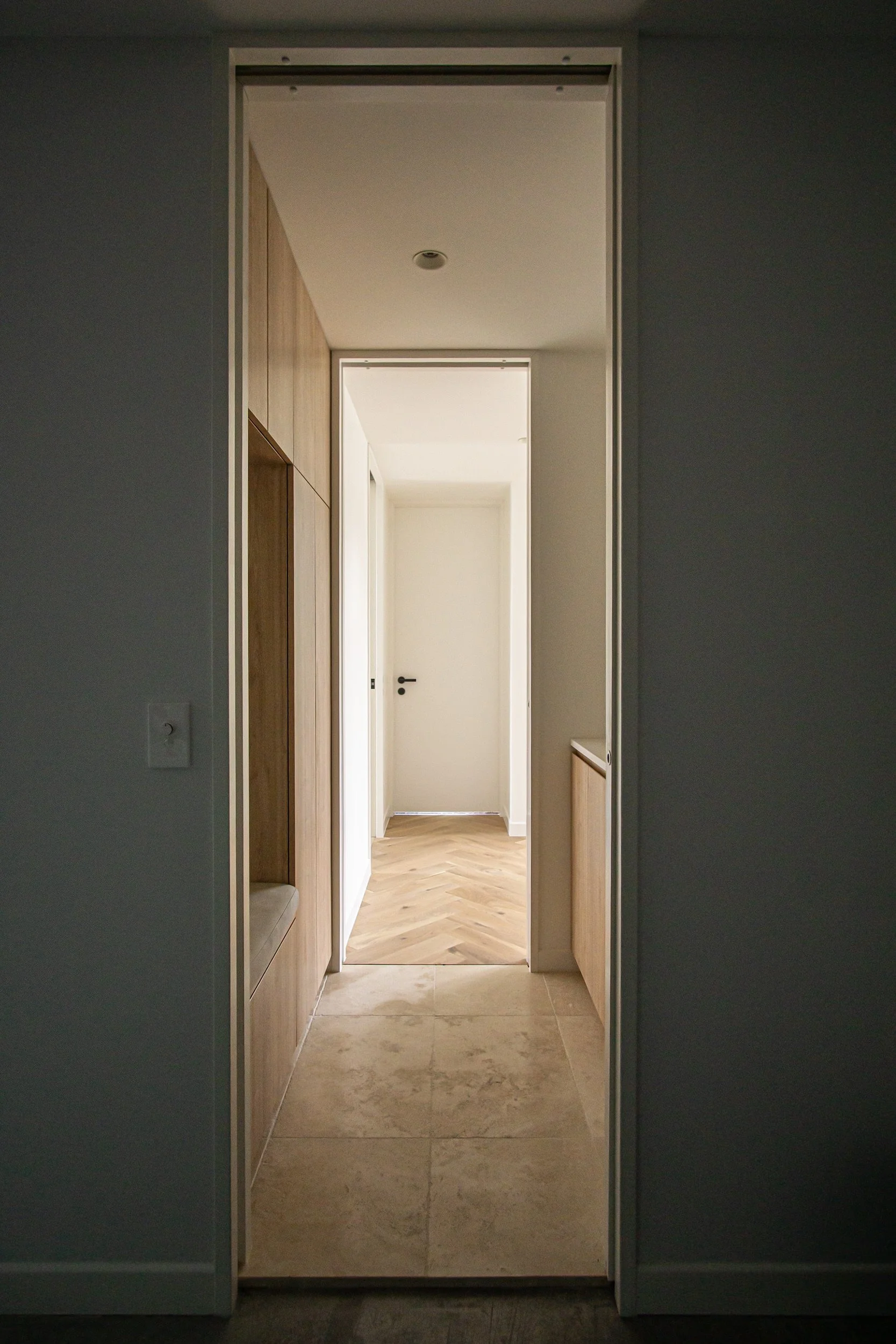 View through an open doorway into a hallway with light-colored walls, wooden cabinets on the left, and a door at the end of the hallway.