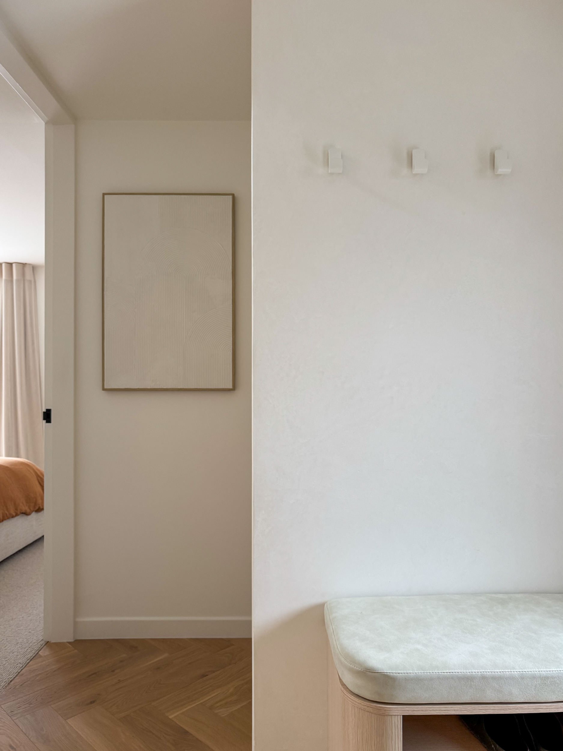 Divided view of a room with beige and white walls, a framed artwork on the beige wall, wooden flooring, a bed with light-colored bedding partially visible, and a white cushioned bench or mattress on the right side.