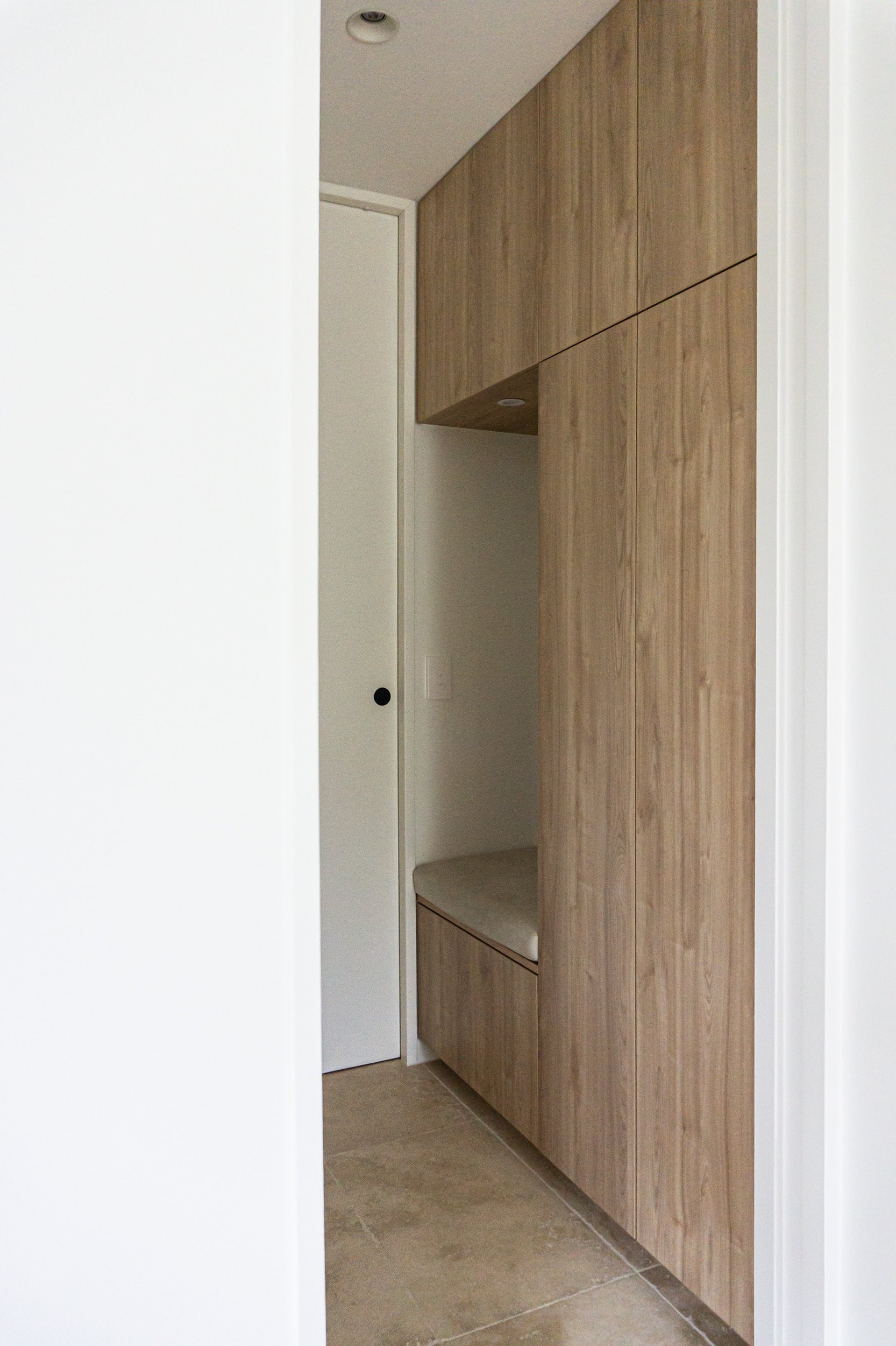 Interior view of a minimalistic space with white walls, a wooden storage unit built into the wall, a small bench, and a tiled floor.