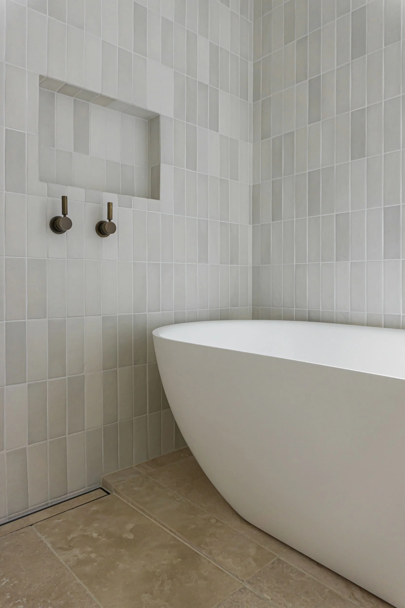 Modern bathroom with a white, freestanding bathtub, beige tiled floor, and walls with small square beige tiles, including a built-in wall shelf and two wall-mounted fixtures.