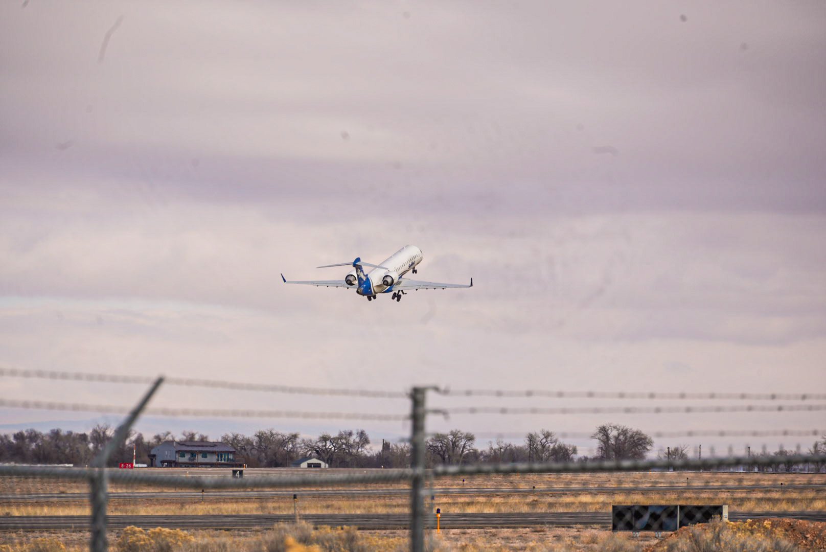 An airplane is taking off from an airport runway with a cloudy sky above and a barbed wire fence in the foreground.