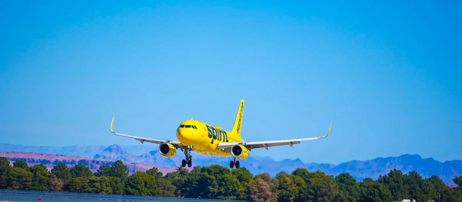 A yellow plane with the logo 'SPIRIT' on it is landing on a runway, with green trees and mountains in the background under a blue sky.