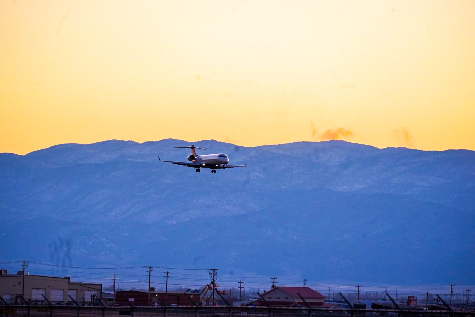 An airplane flying low over a residential area with power lines, with mountains in the background during sunset.