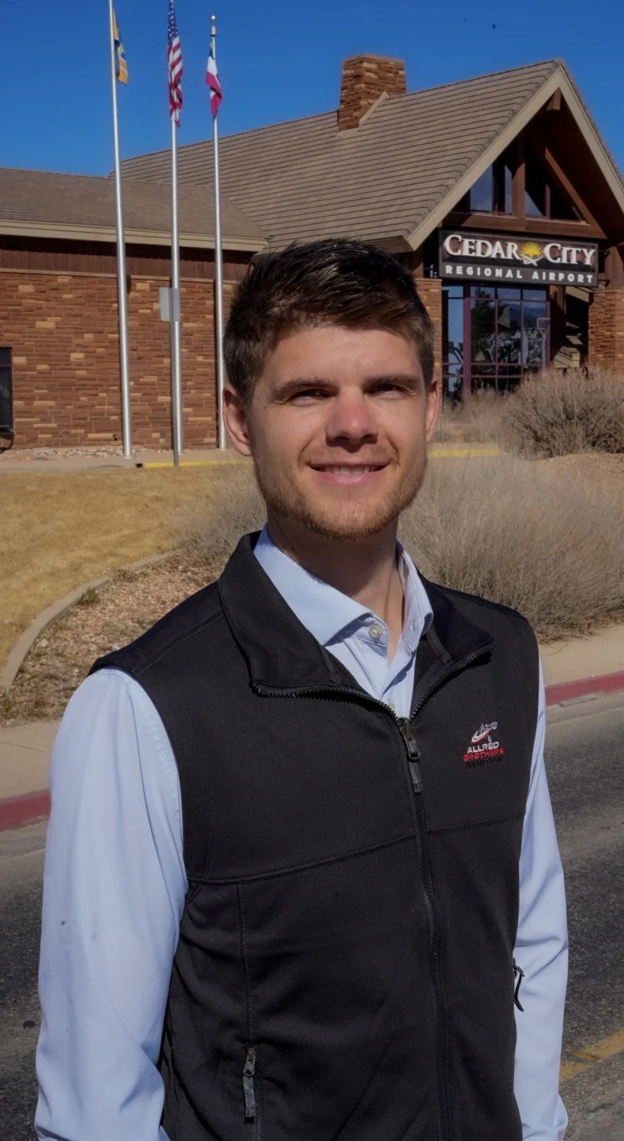 A young man with short brown hair and a beard smiling outdoors in front of Cedar City Regional Airport, with flags and a brick building in the background.