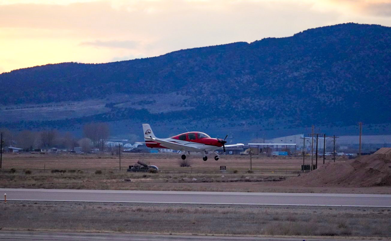 Small red and white airplane taking off or landing on a runway with mountains in the background during sunset or sunrise.