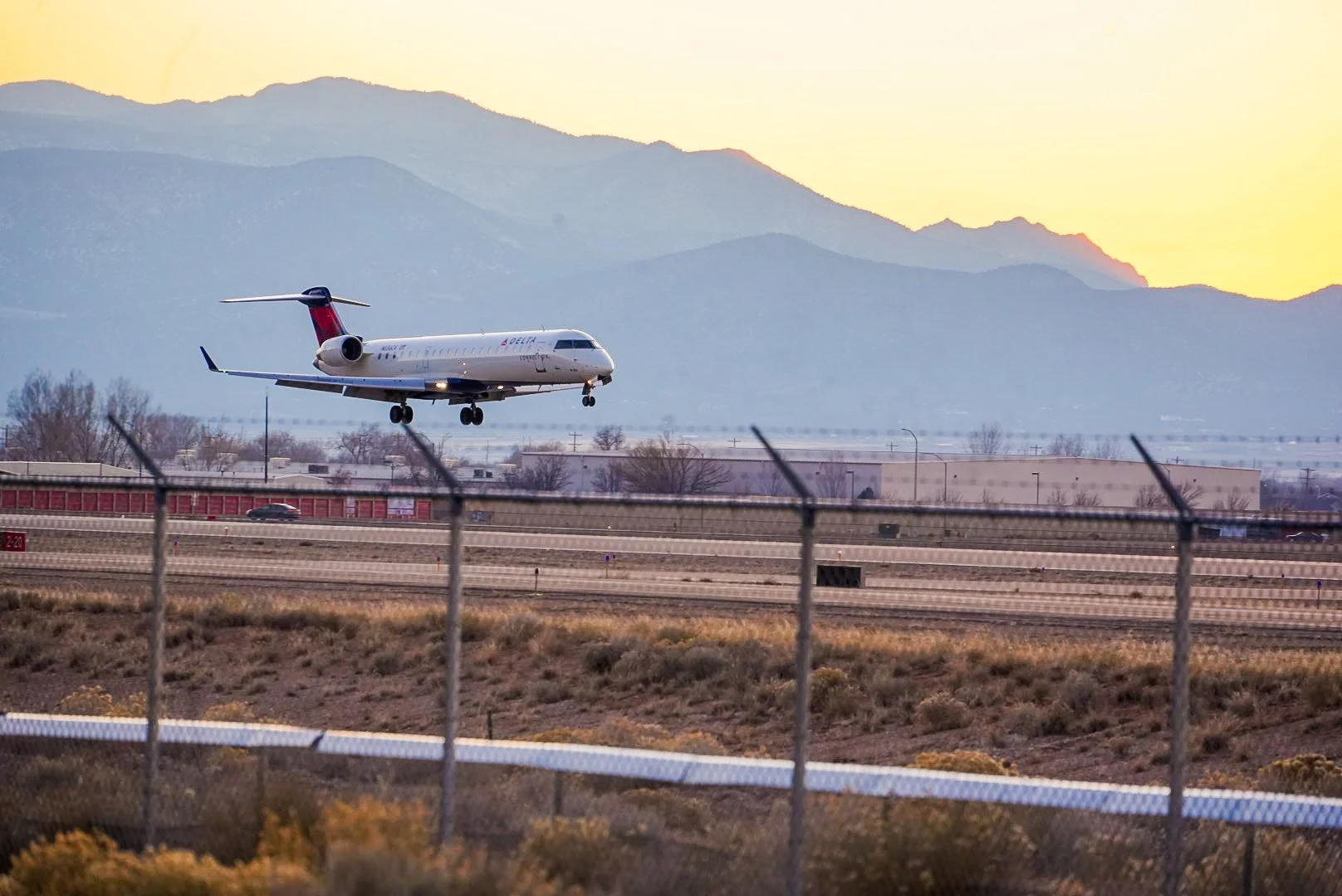 An American Airlines aircraft flying low over the runway during sunset, with mountains in the background and a fenced perimeter in the foreground.