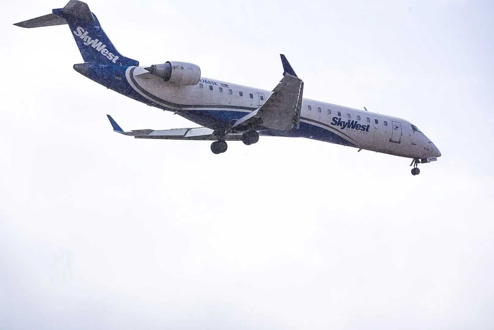 A SkyWest regional jet in flight with landing gear extended, flying through a cloudy sky.