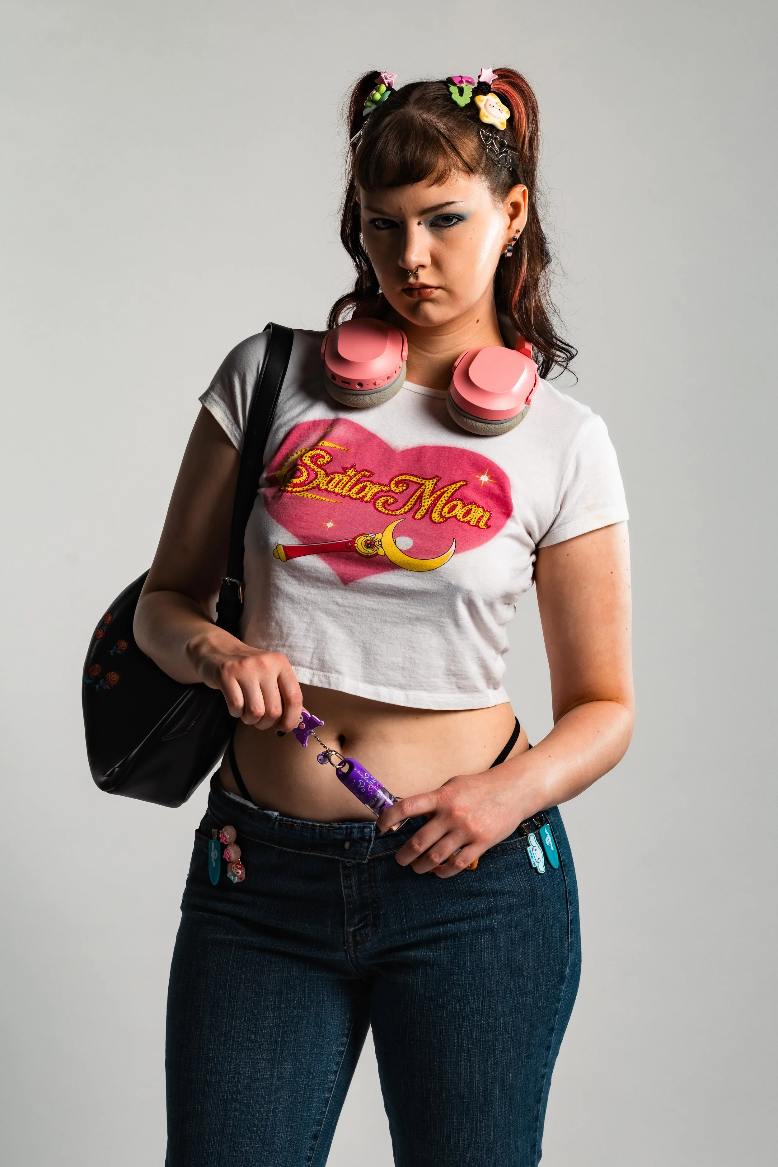 A young woman with dark red hair styled in pigtails, wearing a Sailor Moon T-shirt, dark jeans with character keychains, and pink headphones around her neck. She has bold makeup, multiple earrings, and is holding a purple vape, carrying a black backp