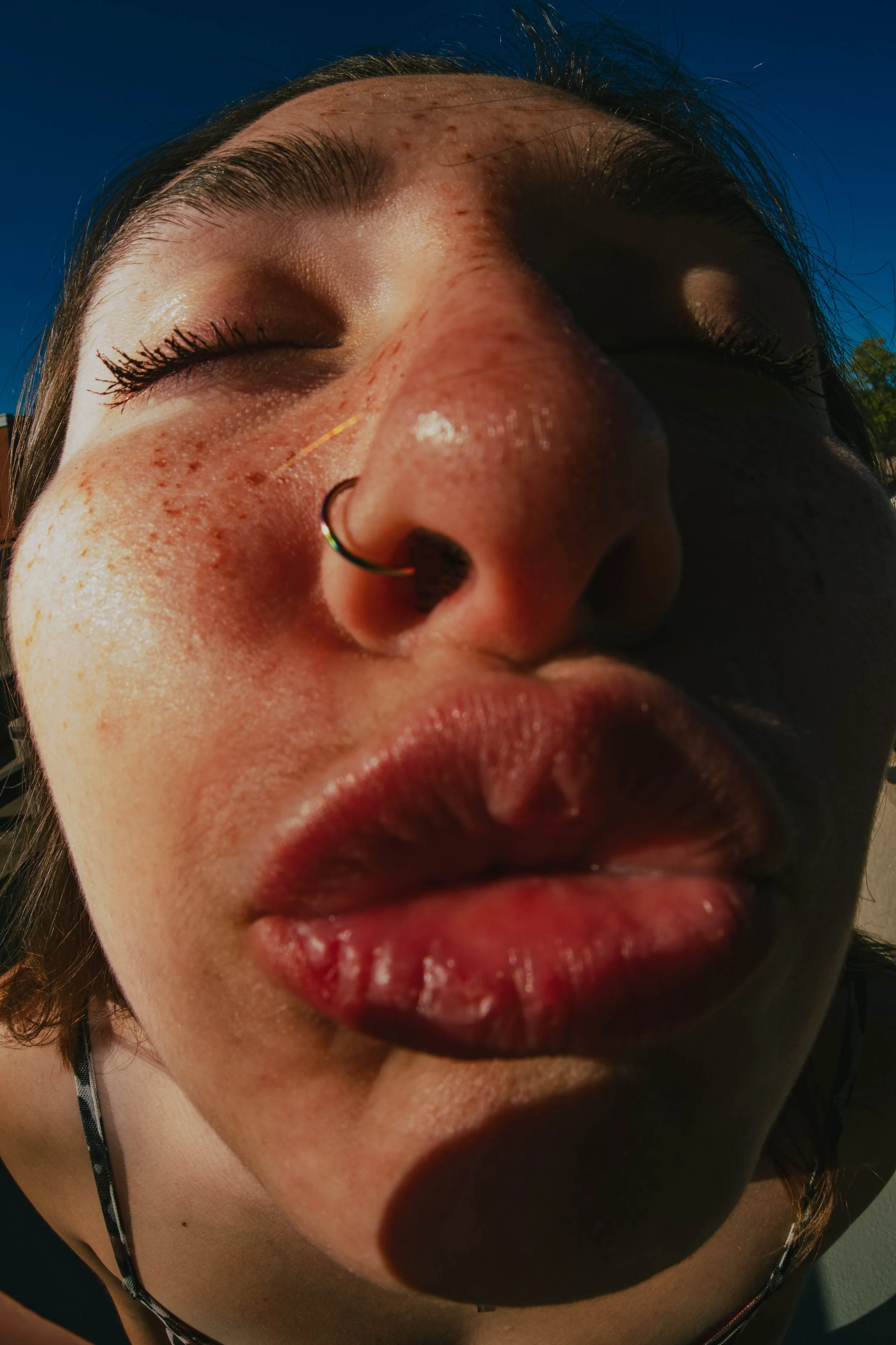 Close-up of a person's face with closed eyes, slightly parted lips, nose piercing, and freckles, taken from a low angle outdoors during daytime.