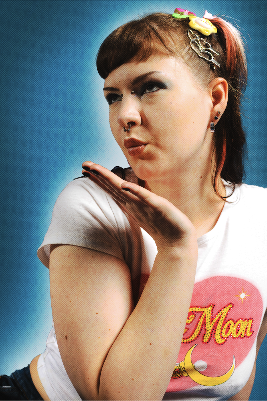 A young woman with colorful hair accessories, piercings, and dark eye makeup is posing with her hand near her face. She is wearing a white t-shirt with a pink heart and the word 'Moon' on it and has a blue background.